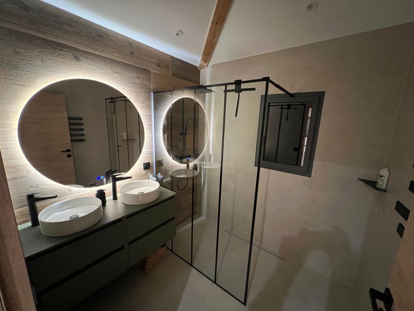 Salle de bains moderne avec deux lavabos, une douche en verre et un grand miroir rond à cadre lumineux.