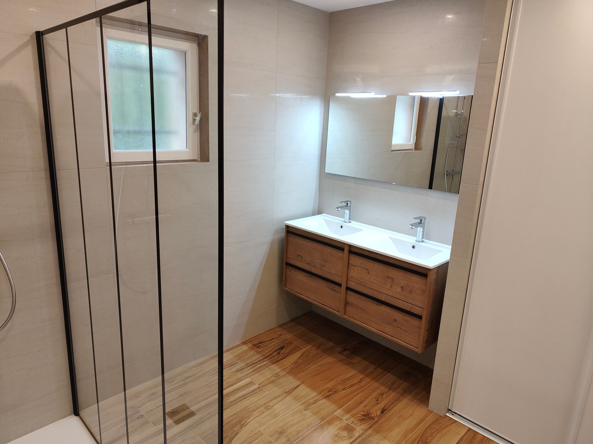 Salle de bains moderne avec sol en imitation bois, murs gris clair, douche en verre et meuble-lavabo à double vasque.