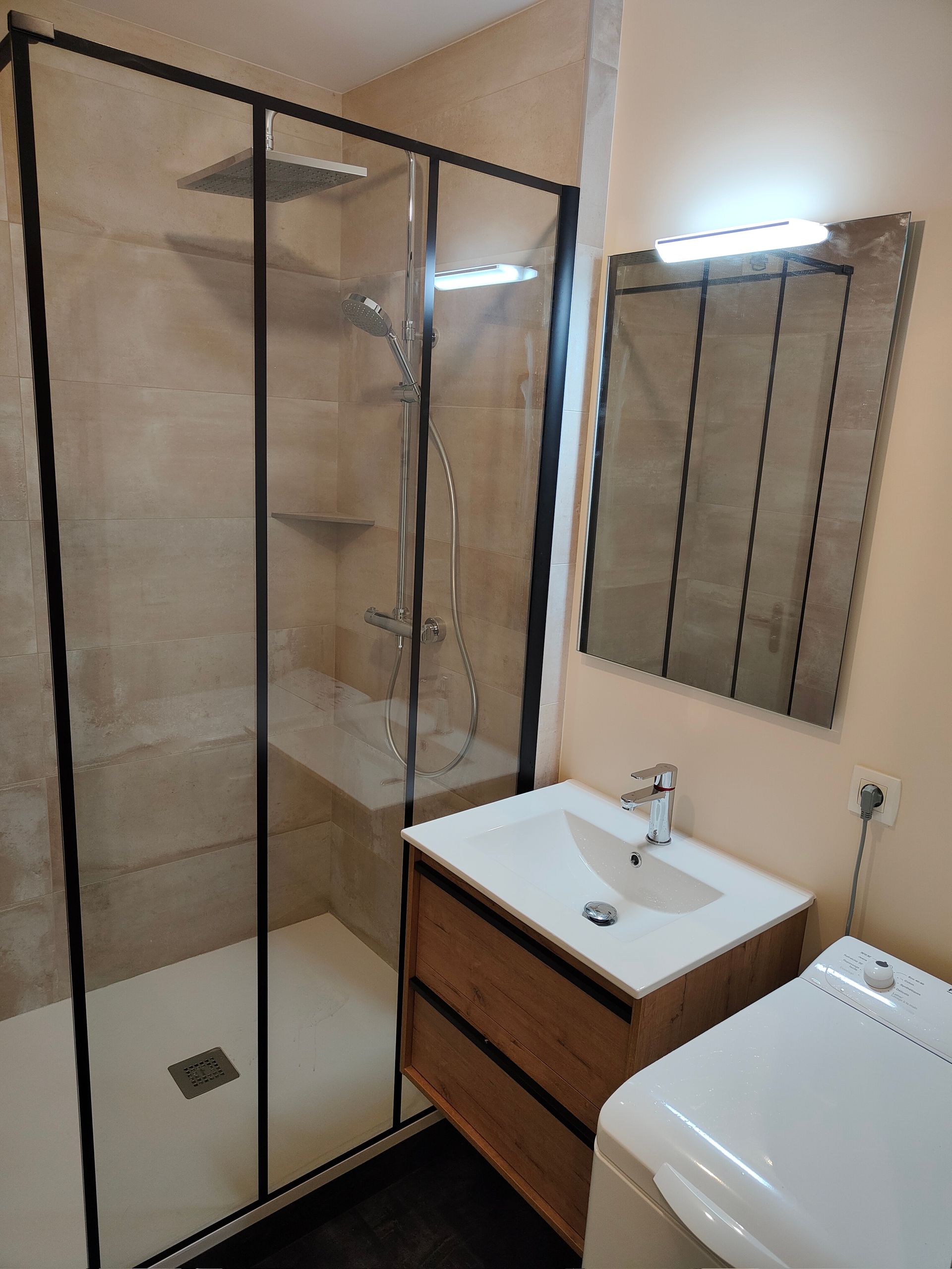 Salle de bains avec douche, meuble-lavabo et miroir ; carrelage beige, cadre de douche noir et lavabo blanc.
