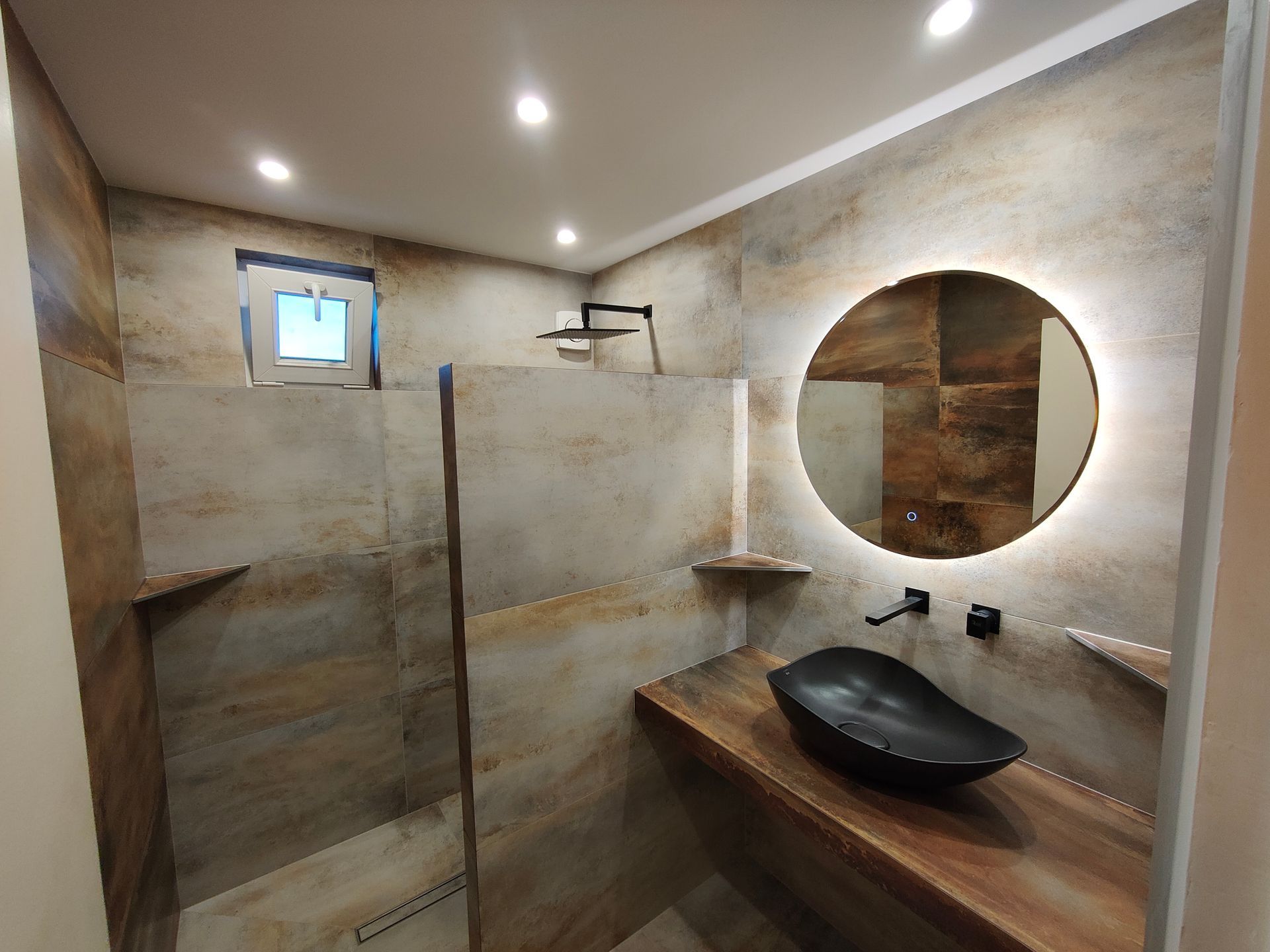 Salle de bains rustique avec murs carrelés bruns et gris, vasque noire et miroir rond éclairé.