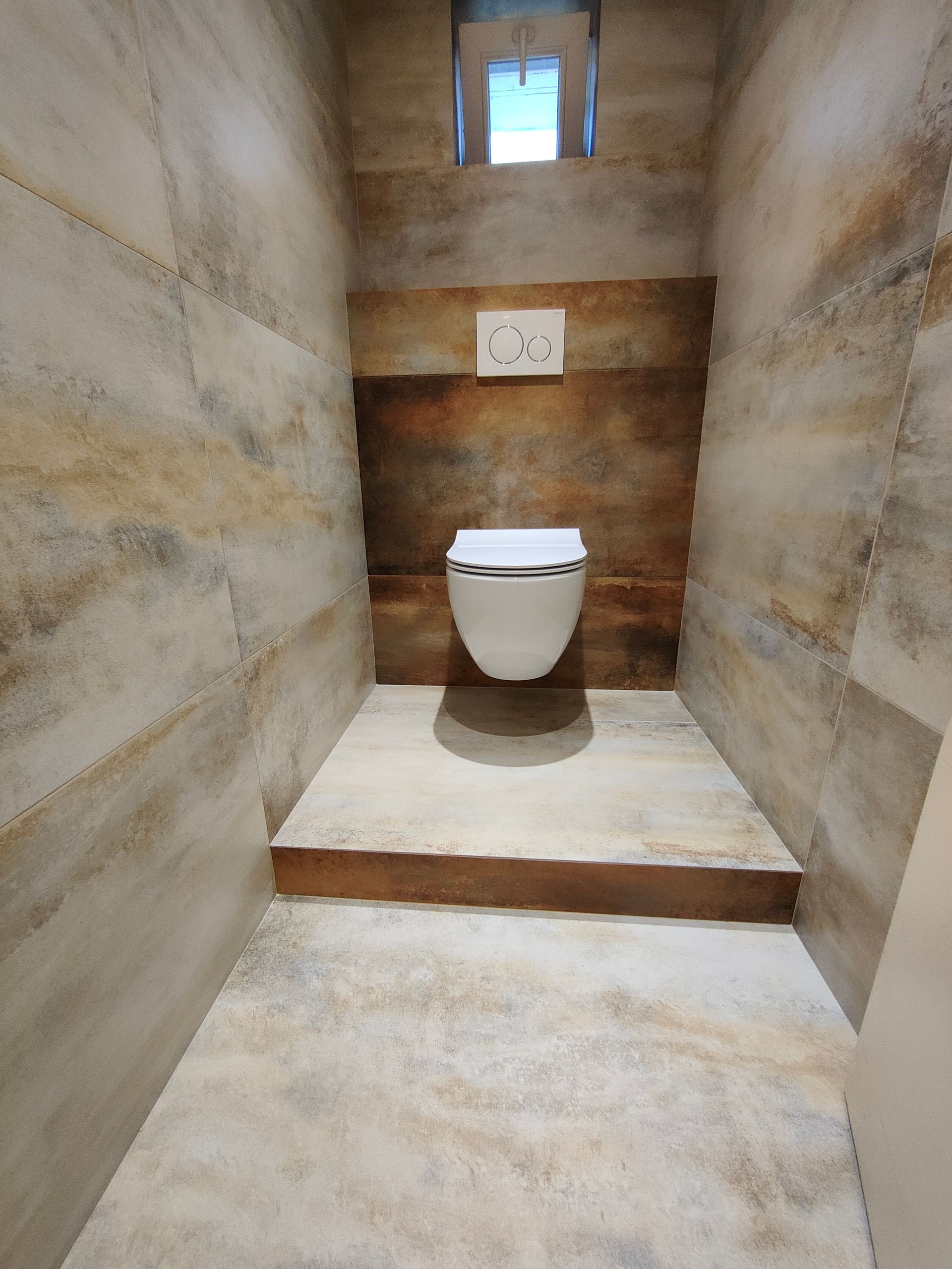 Une petite salle de bains moderne avec des toilettes suspendues. Murs couleur pierre beige et marron. Petite fenêtre au plafond.