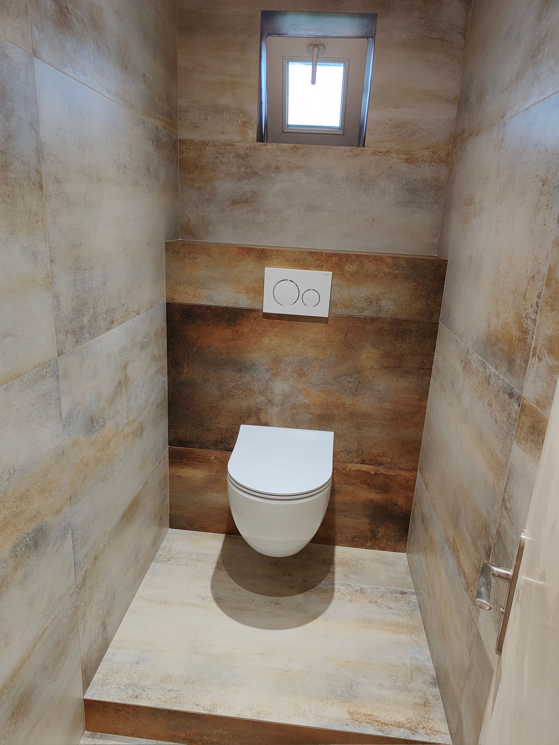 Petite cabine de toilettes fermée, carrelée de faïence beige et marron imitation pierre. Les toilettes sont fixées au mur, sous le bouton de chasse d'eau.