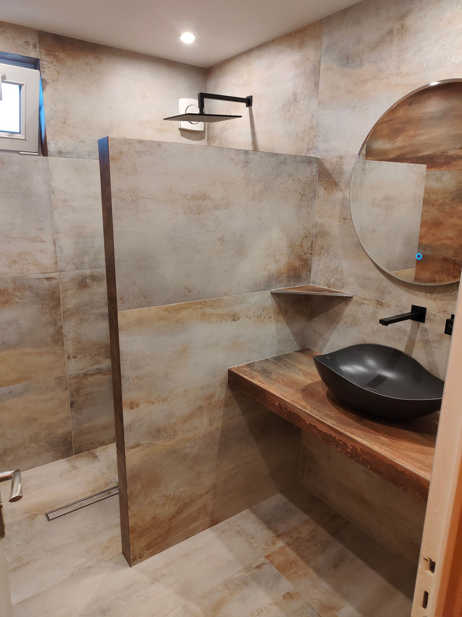 Salle de bains avec douche, lavabo et grand miroir rond. Tons naturels, carrelage en pierre et robinetterie noire.