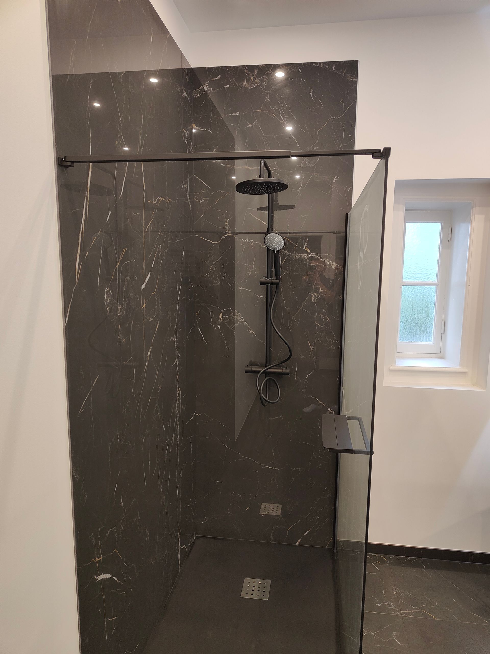 Cabine de douche avec carrelage imitation marbre gris foncé, robinetterie noire et petite fenêtre.