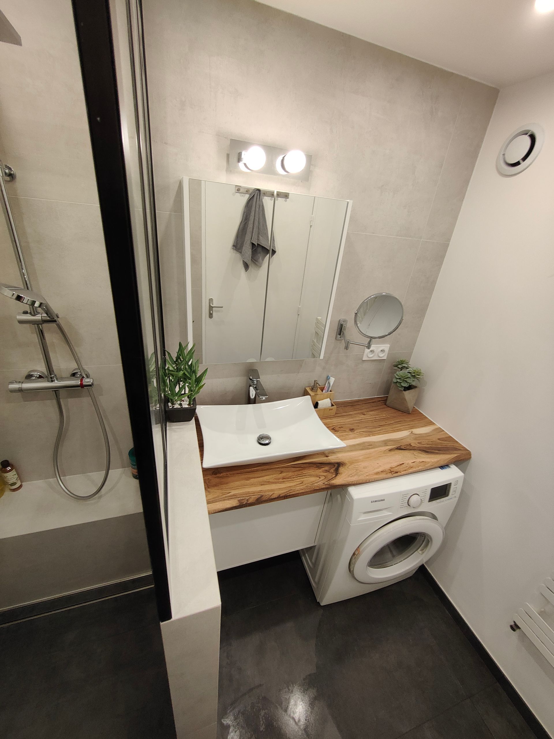 Salle de bains moderne avec lave-linge, plan de travail en bois et cabine de douche en verre.