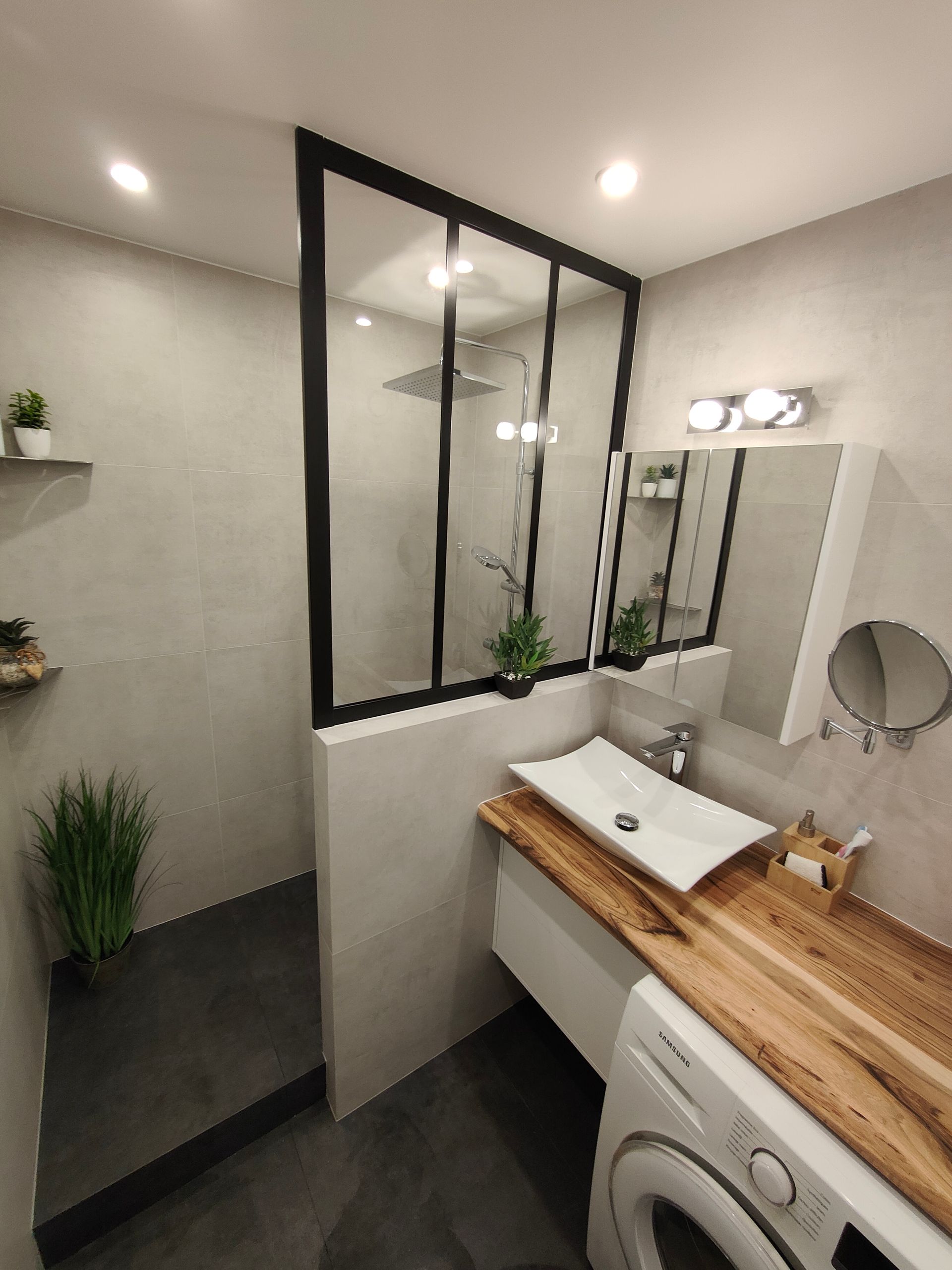 Salle de bains avec douche à l'italienne, paroi de douche en verre à cadre noir et comptoir en bois avec lavabo. Murs gris et lave-linge.