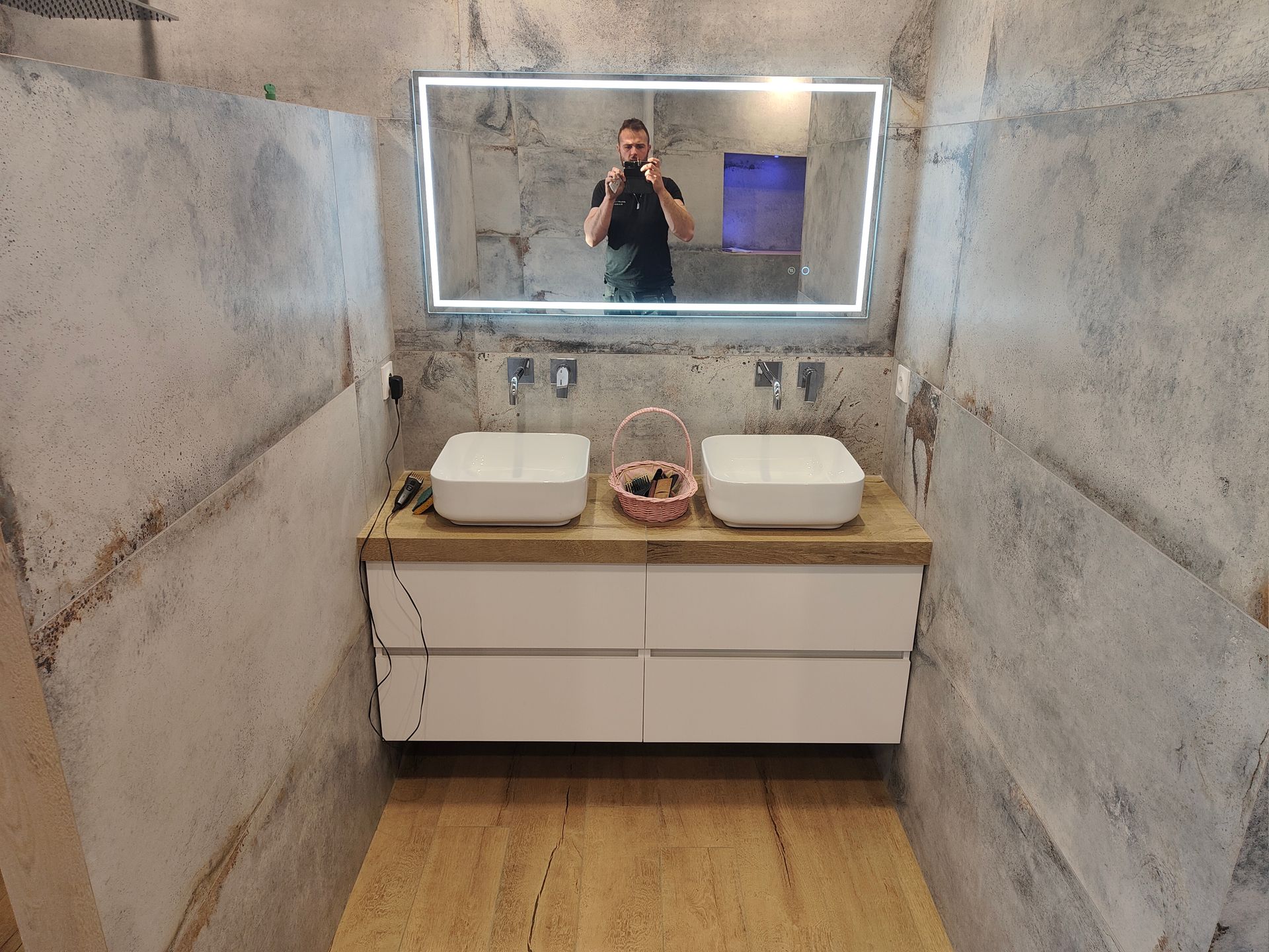 Salle de bains avec deux lavabos blancs, un grand miroir et une personne qui se fait photographier dedans.