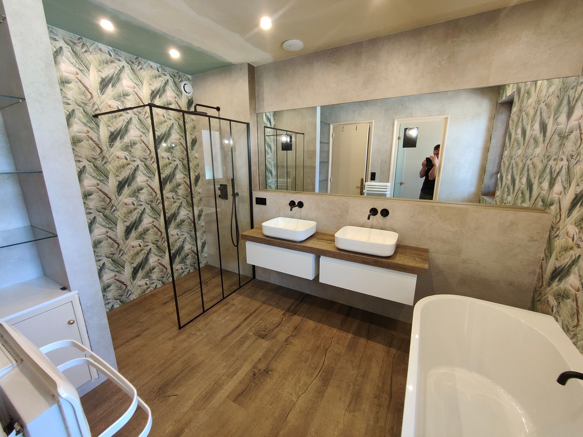 Salle de bains avec parquet, double vasque blanche, douche en verre et baignoire ; papier peint à motifs de feuilles vertes.