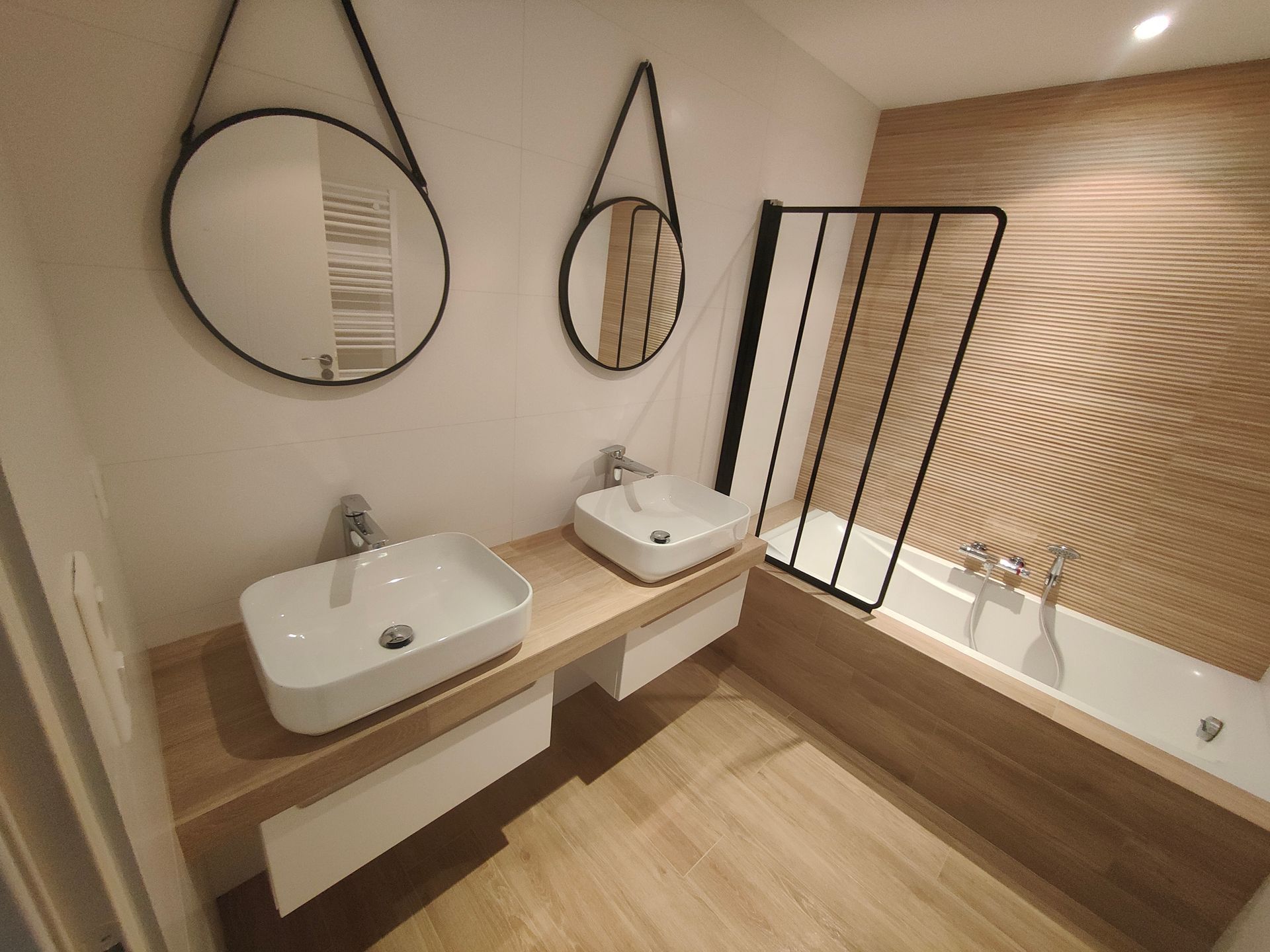 Salle de bains moderne avec deux lavabos, des miroirs et une baignoire avec une paroi de douche en verre à cadre noir, et des éléments en bois.