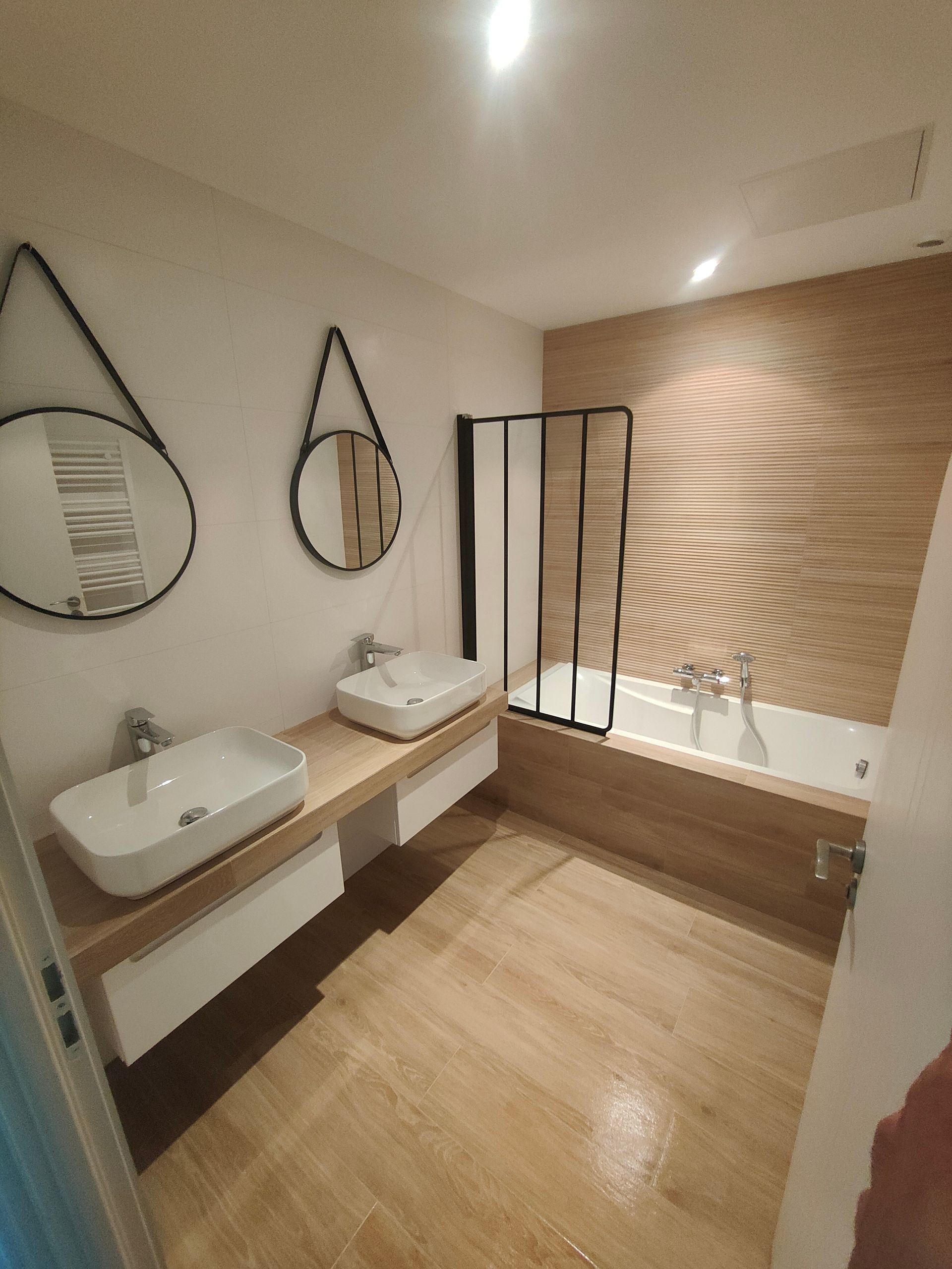 Salle de bains avec des touches de bois et de blanc : deux lavabos avec miroirs, baignoire avec paroi noire.