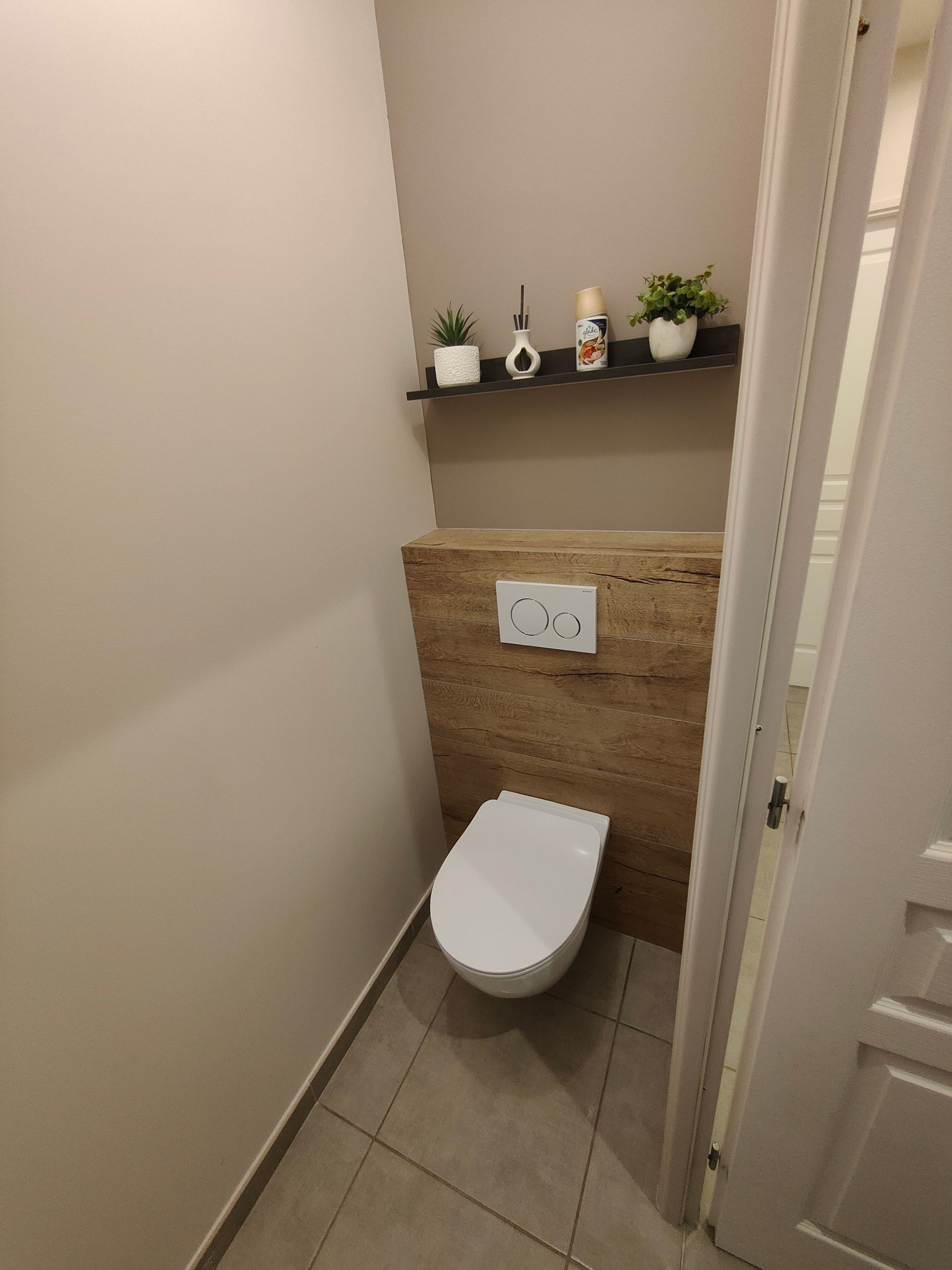 Petite salle de bains avec toilettes et étagère ornée de plantes et d'objets décoratifs ; murs beiges, sol carrelé gris et porte blanche.