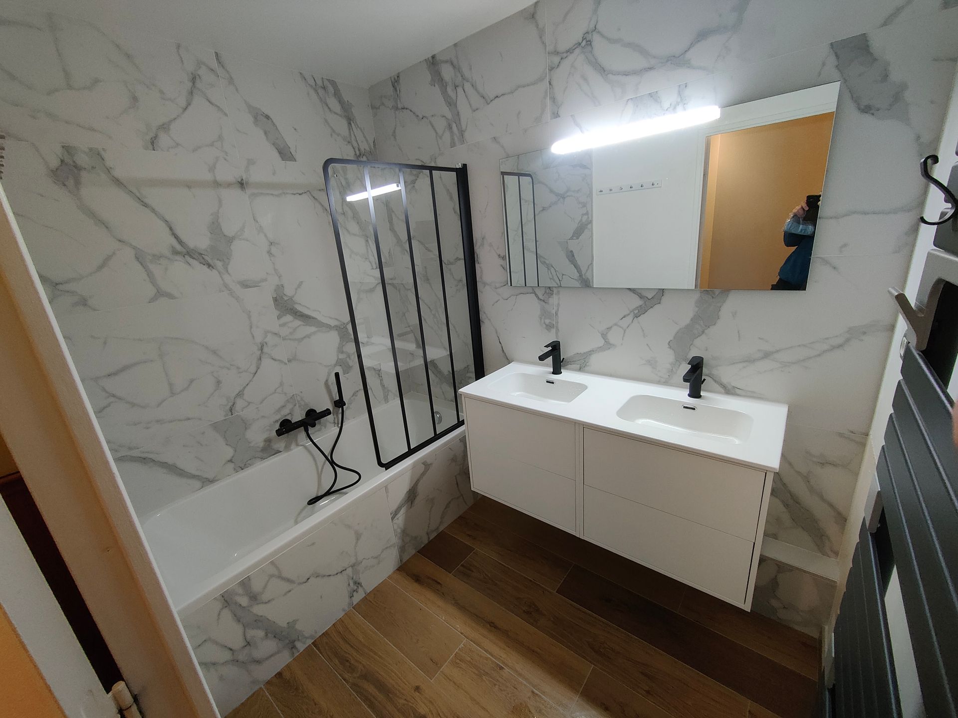 Salle de bains moderne avec carrelage en marbre blanc, robinetterie noire et sol imitation bois.