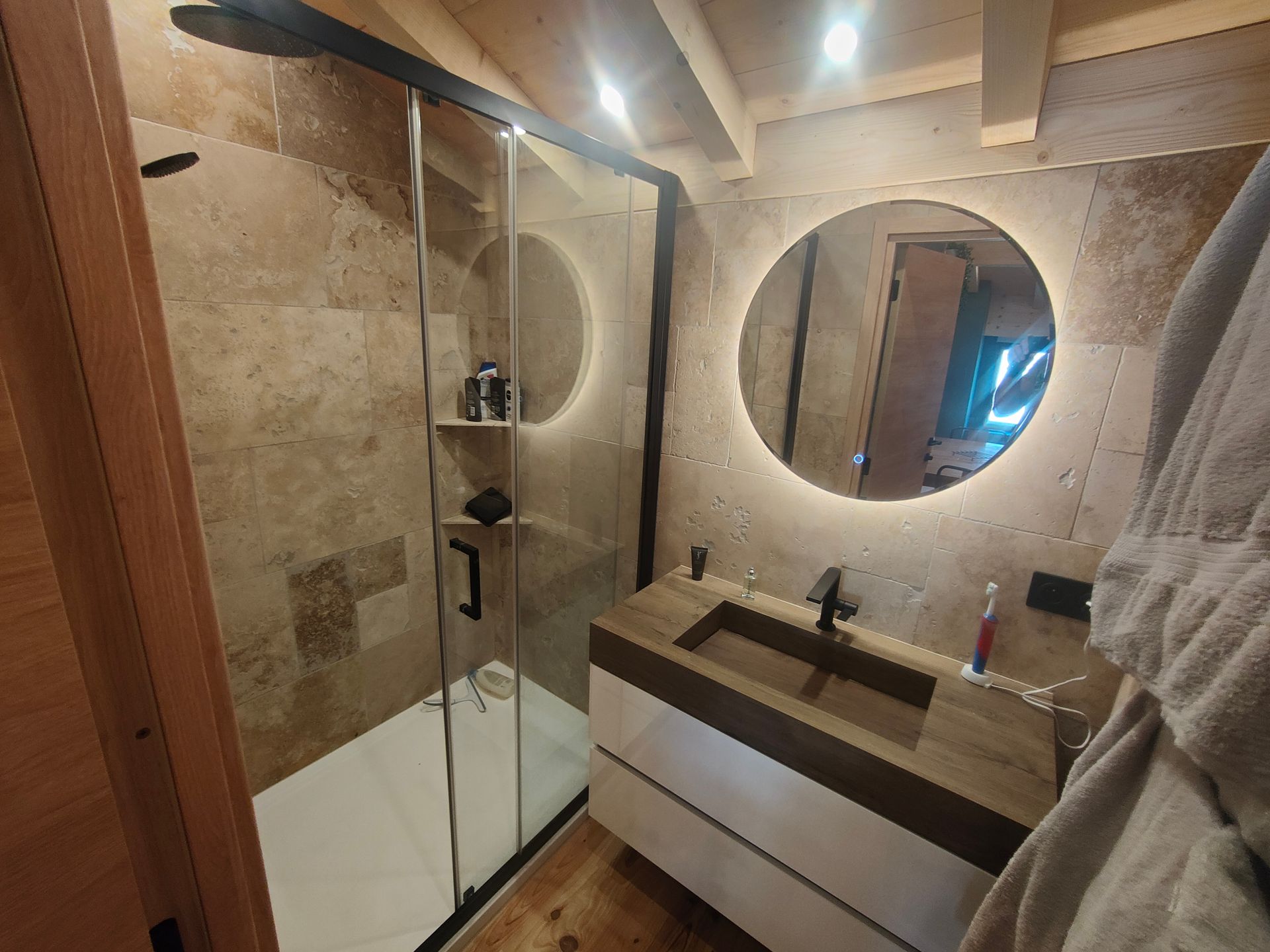 Petite salle de bains moderne avec douche, lavabo, miroir rond et éléments en bois.