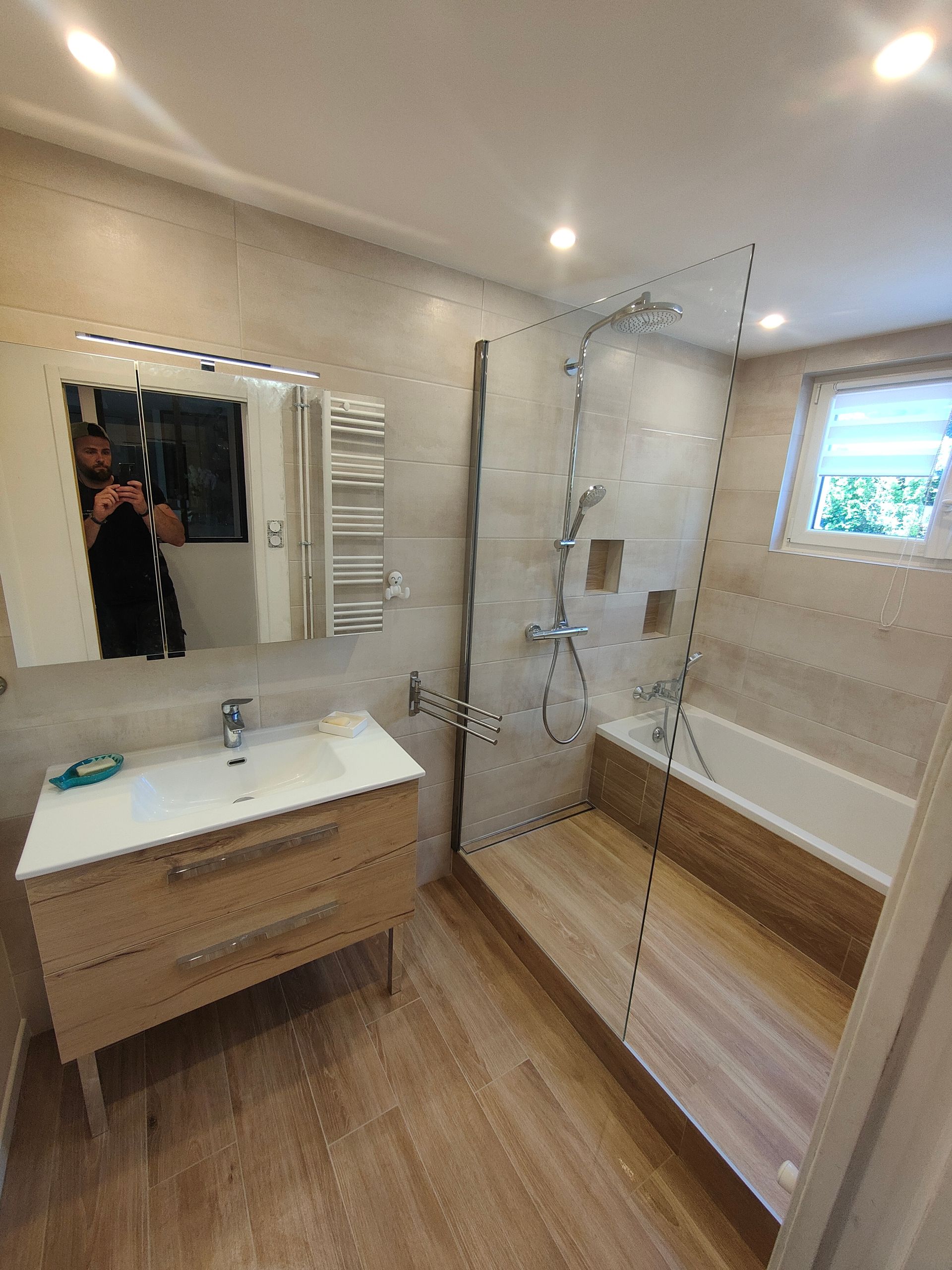 Salle de bains carrelée avec carrelage imitation bois. Baignoire-douche avec paroi de verre. Meuble-lavabo en bois clair, miroir et petite fenêtre.
