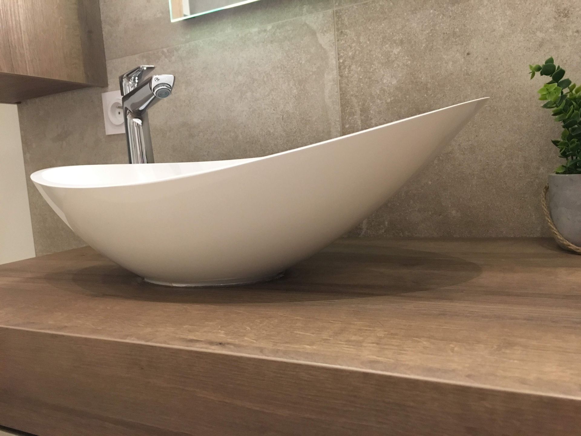 Lavabo blanc en forme de bateau sur un plan de travail en bois avec robinet chromé. Mur beige et petite plante en pot.