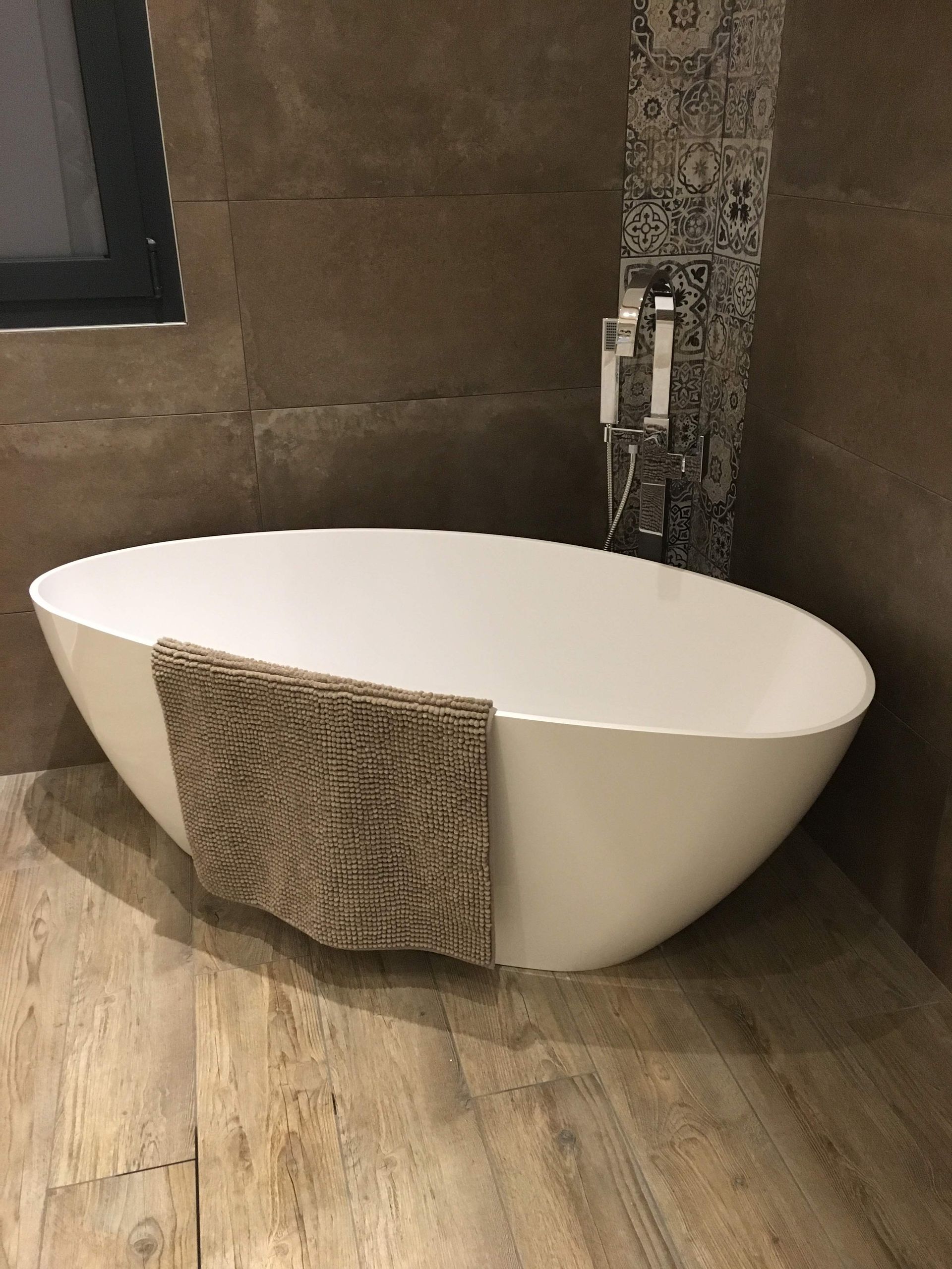 Baignoire ovale blanche avec un tapis marron, dans une salle de bains aux murs carrelés de marron et au sol imitation bois.