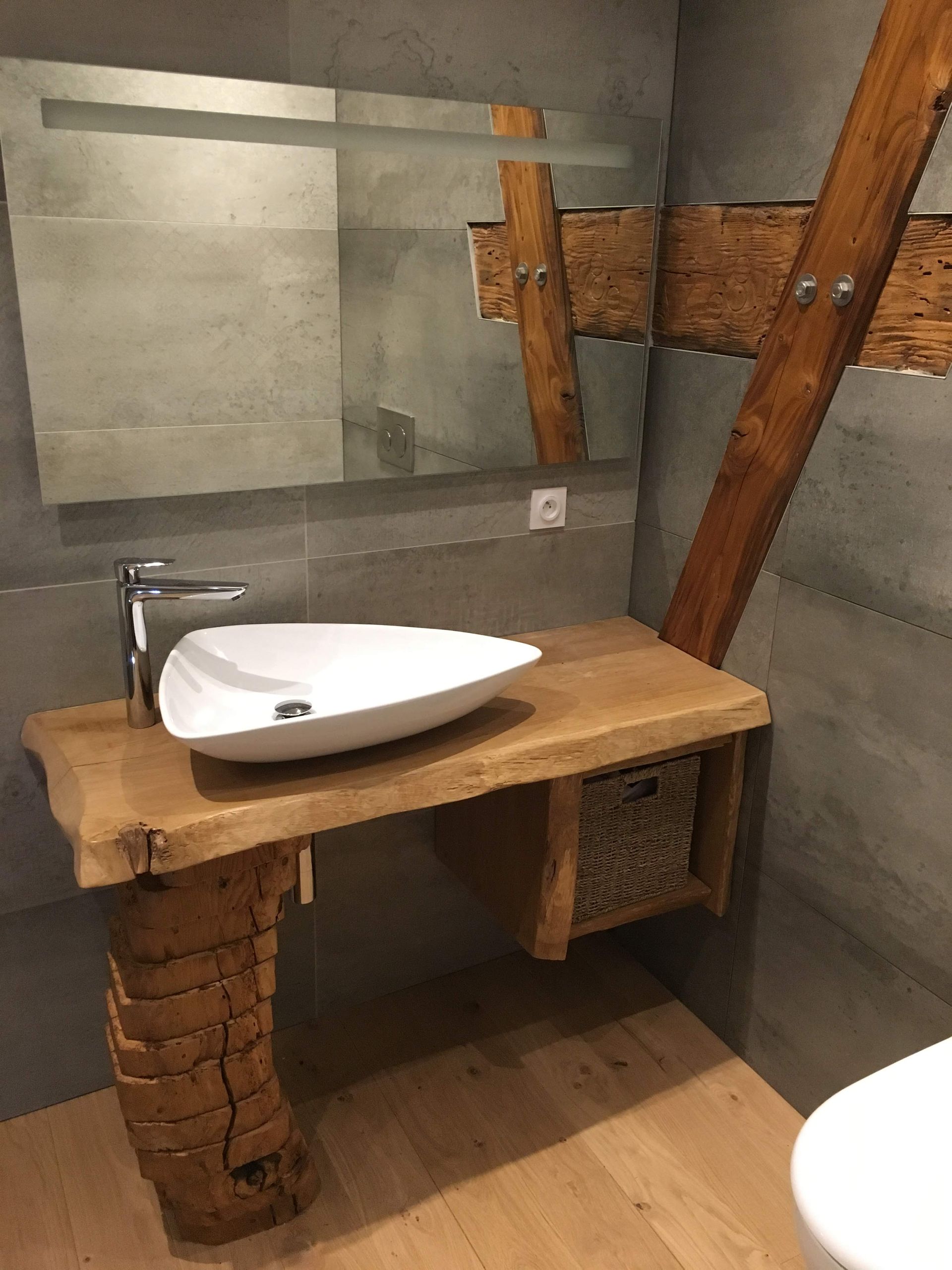 Meuble-lavabo rustique en bois avec vasque dans une salle de bains. Murs carrelés gris, grand miroir et poutres apparentes.