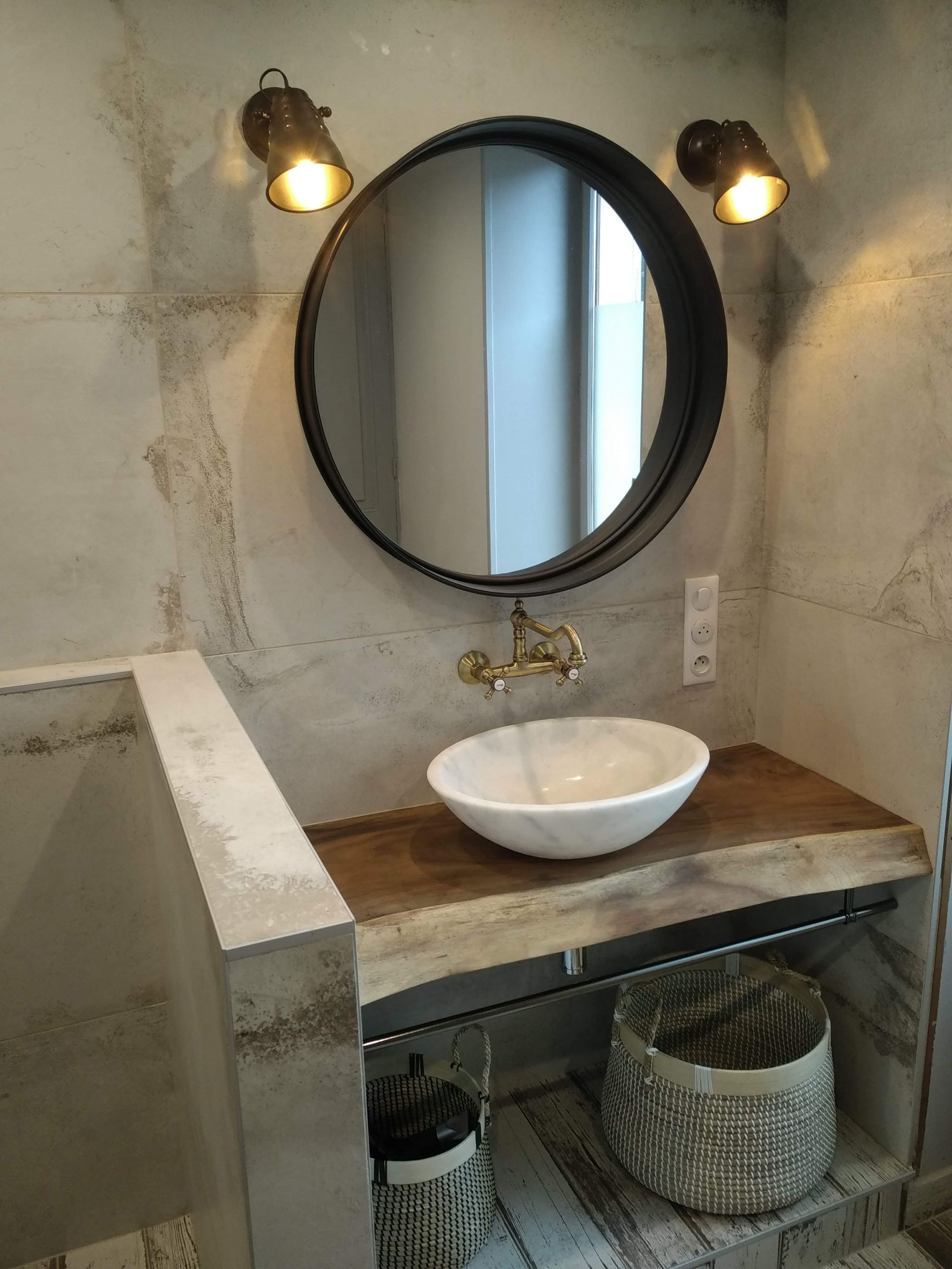 Salle de bains avec miroir rond, lavabo en marbre sur bois, robinetterie en laiton et paniers tressés.