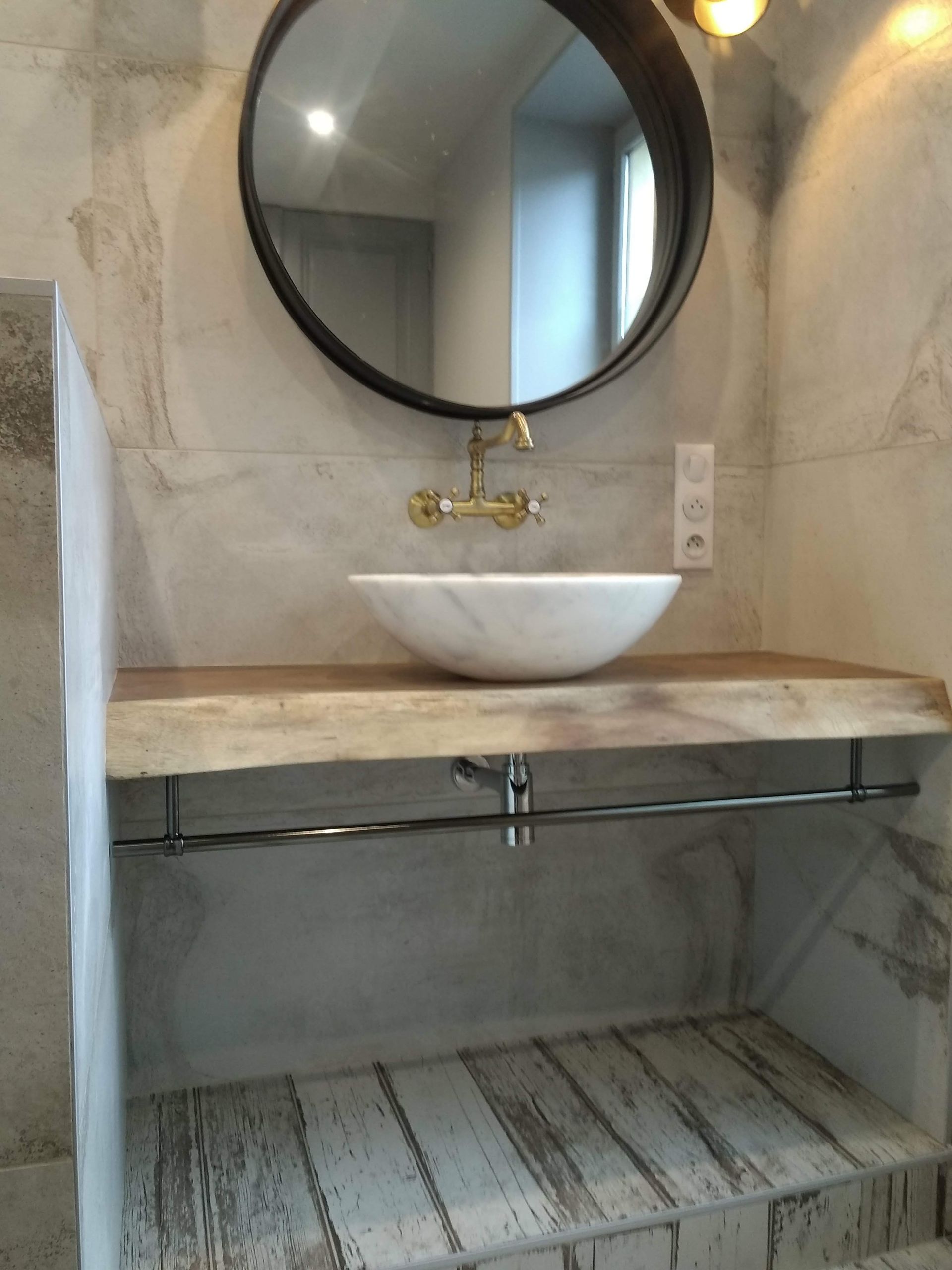 Salle de bains avec vasque en marbre, robinet en laiton, miroir rond et étagère en bois.