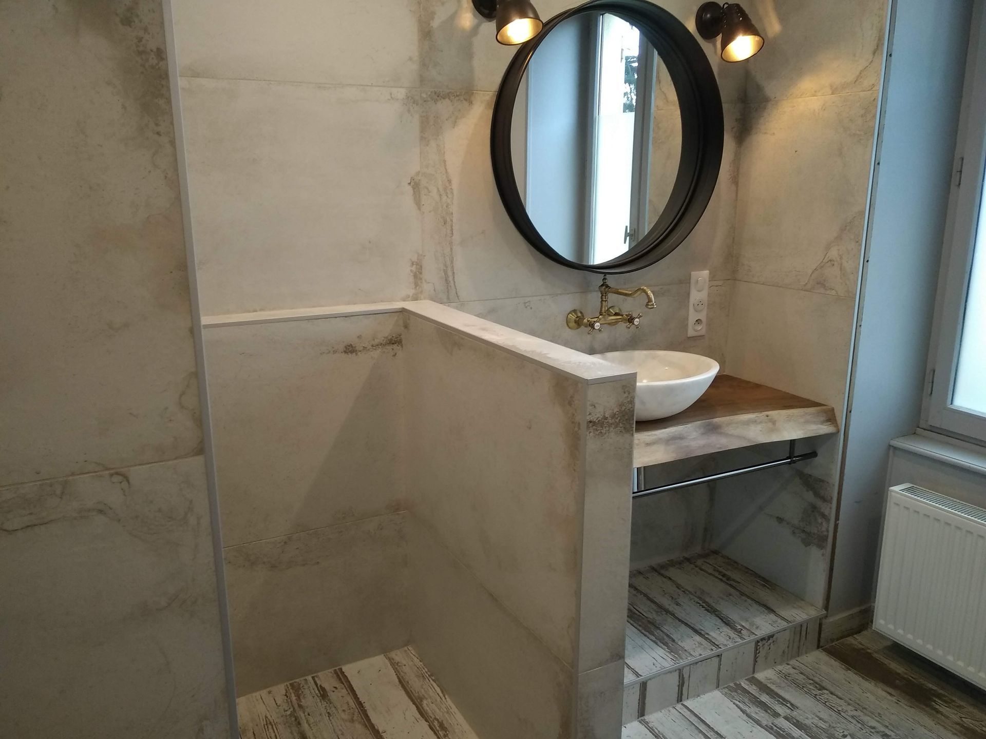 Salle de bains avec miroir ovale, robinet doré et lavabo suspendu. Murs clairs et plan de travail en bois.