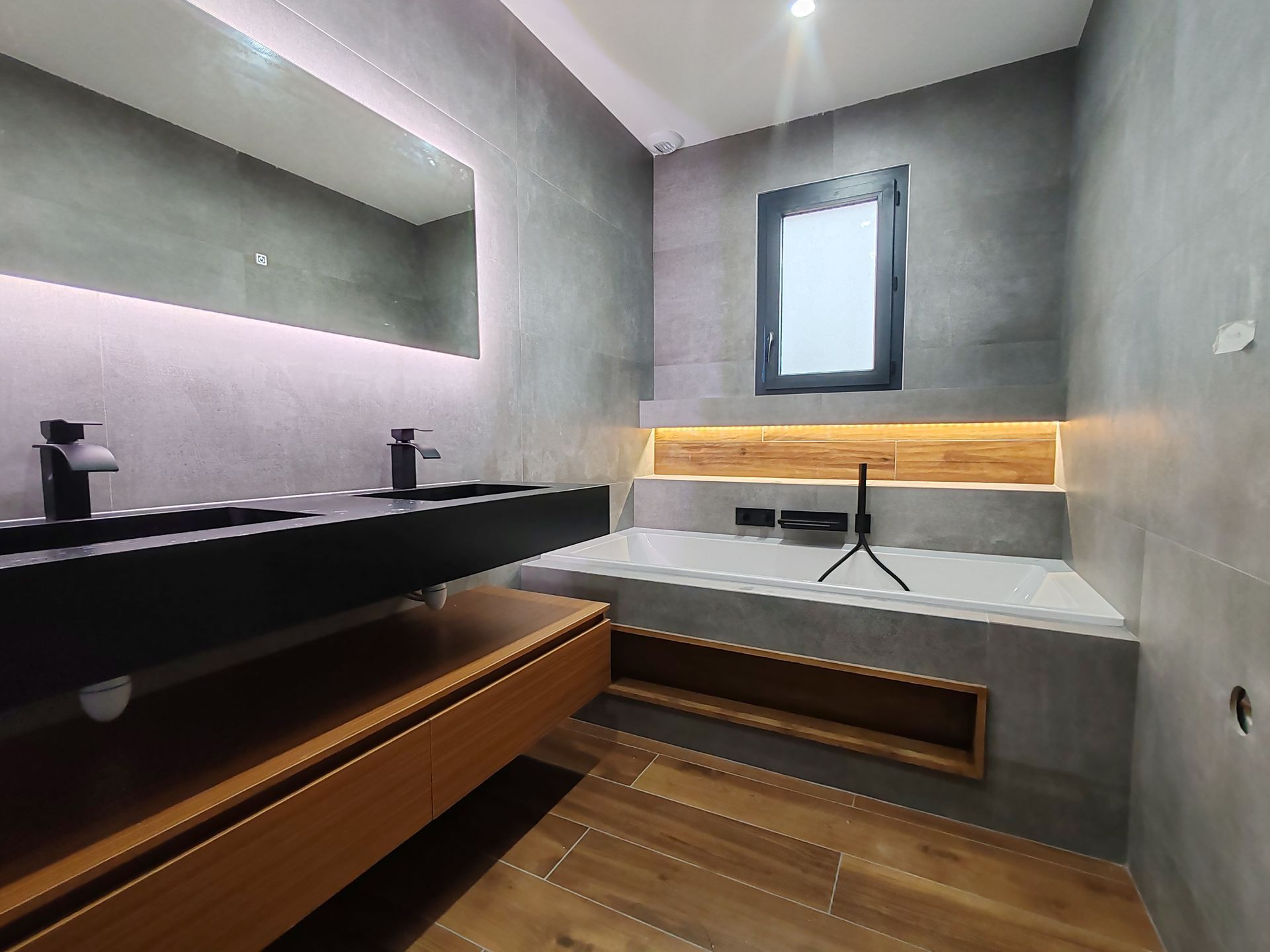 Salle de bains moderne avec murs carrelés gris, lavabos noirs, meuble-lavabo en bois et baignoire encastrée.