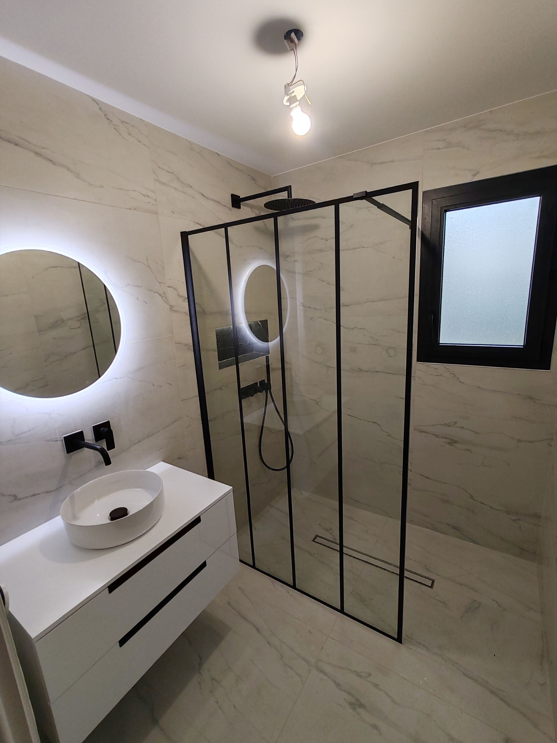 Salle de bains moderne avec douche en verre, miroir rond, meuble-lavabo blanc et petite fenêtre dépolie.