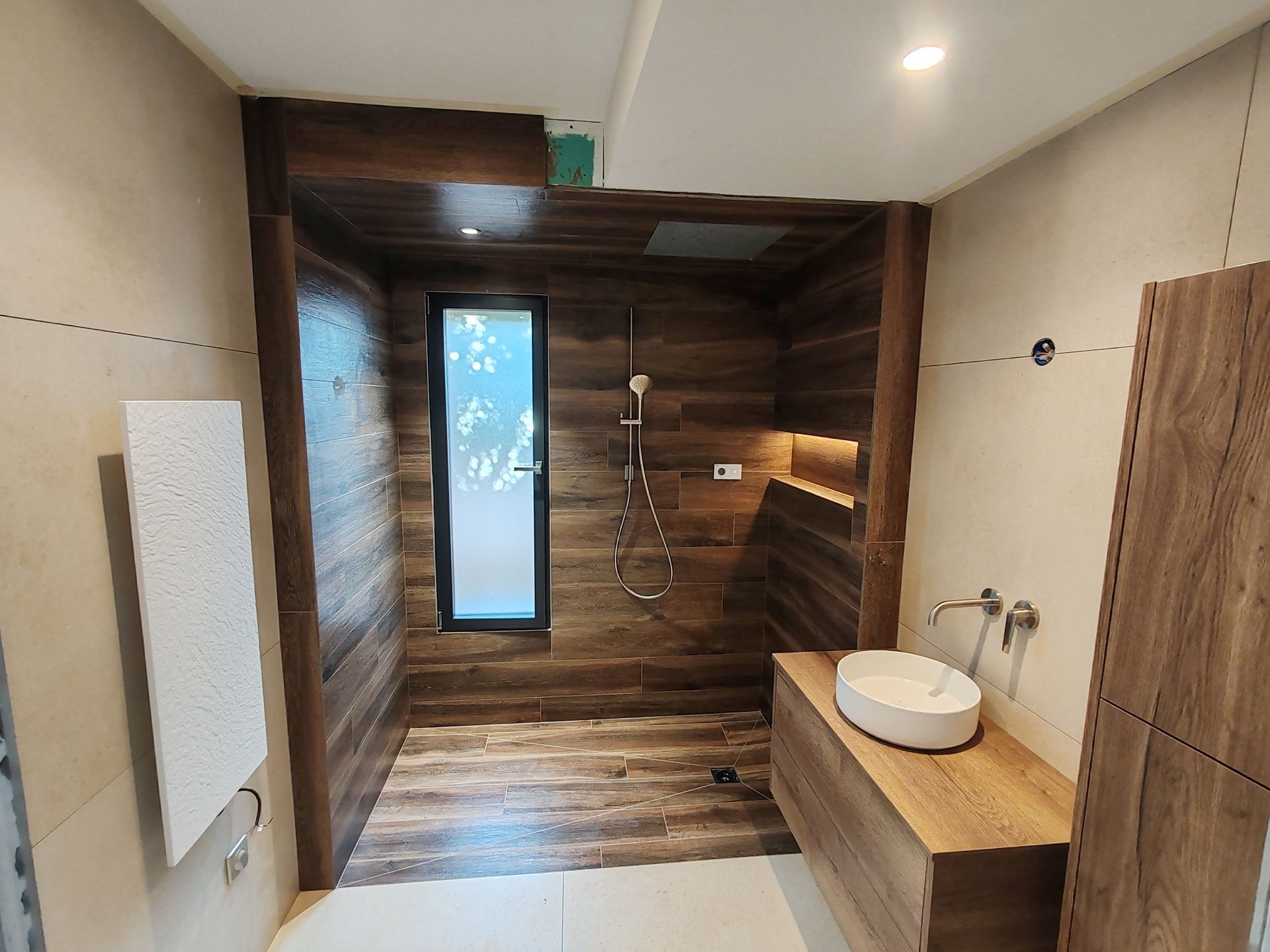 Salle de bains avec carrelage imitation bois marron, douche, fenêtre et vasque blanche sur un meuble-lavabo en bois.
