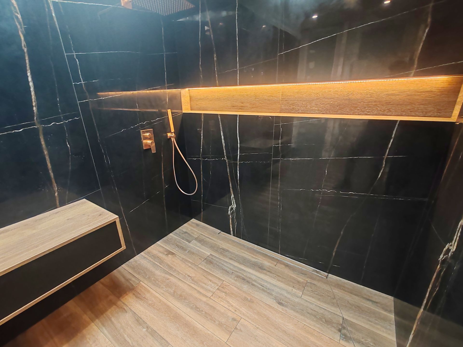 Douche carrelée foncée avec banc imitation bois et robinetterie dorée. Éclairage d'ambiance doré.