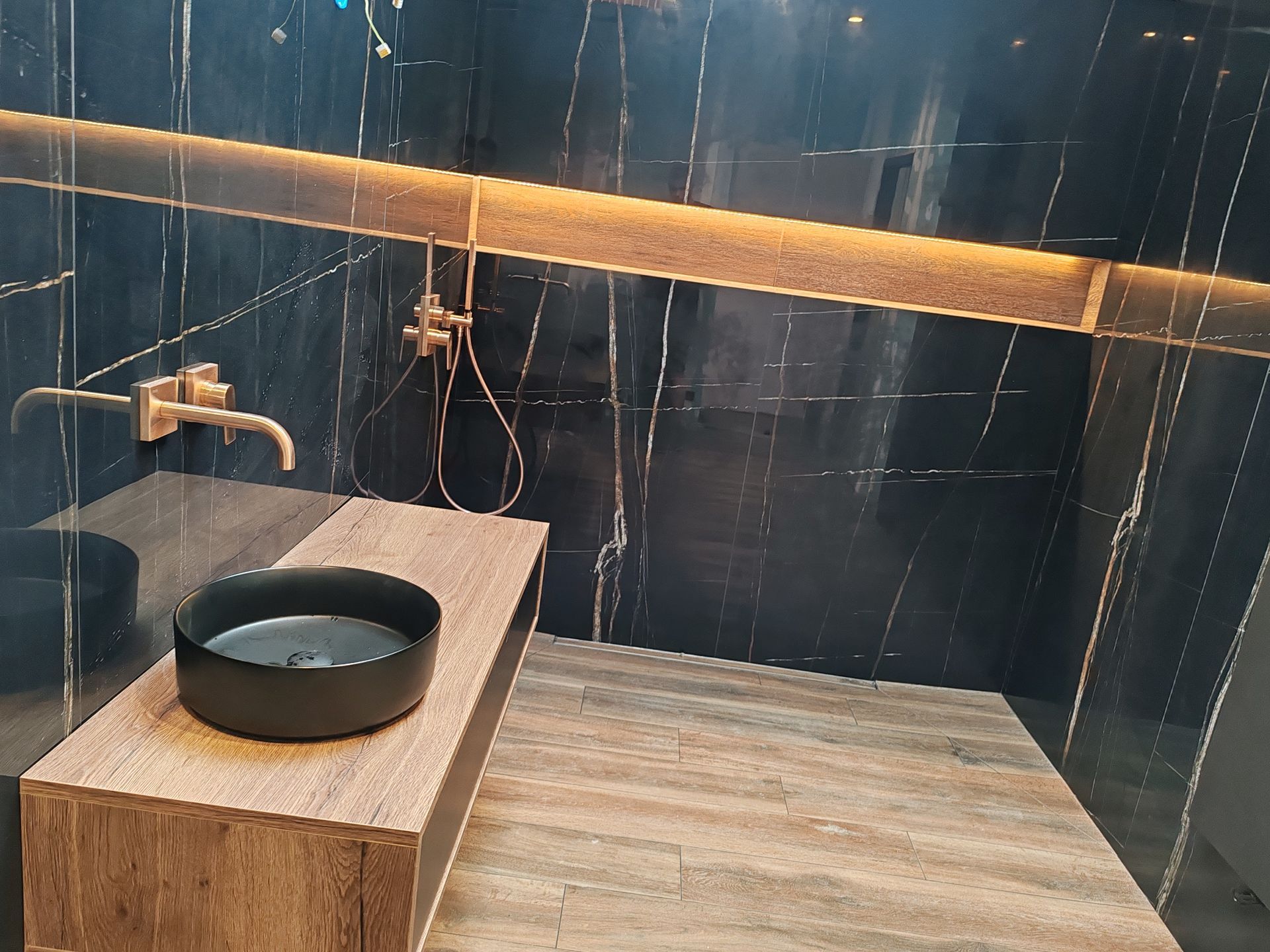 Salle de bains avec murs en marbre noir, comptoir en bois, lavabo noir et robinetterie dorée.