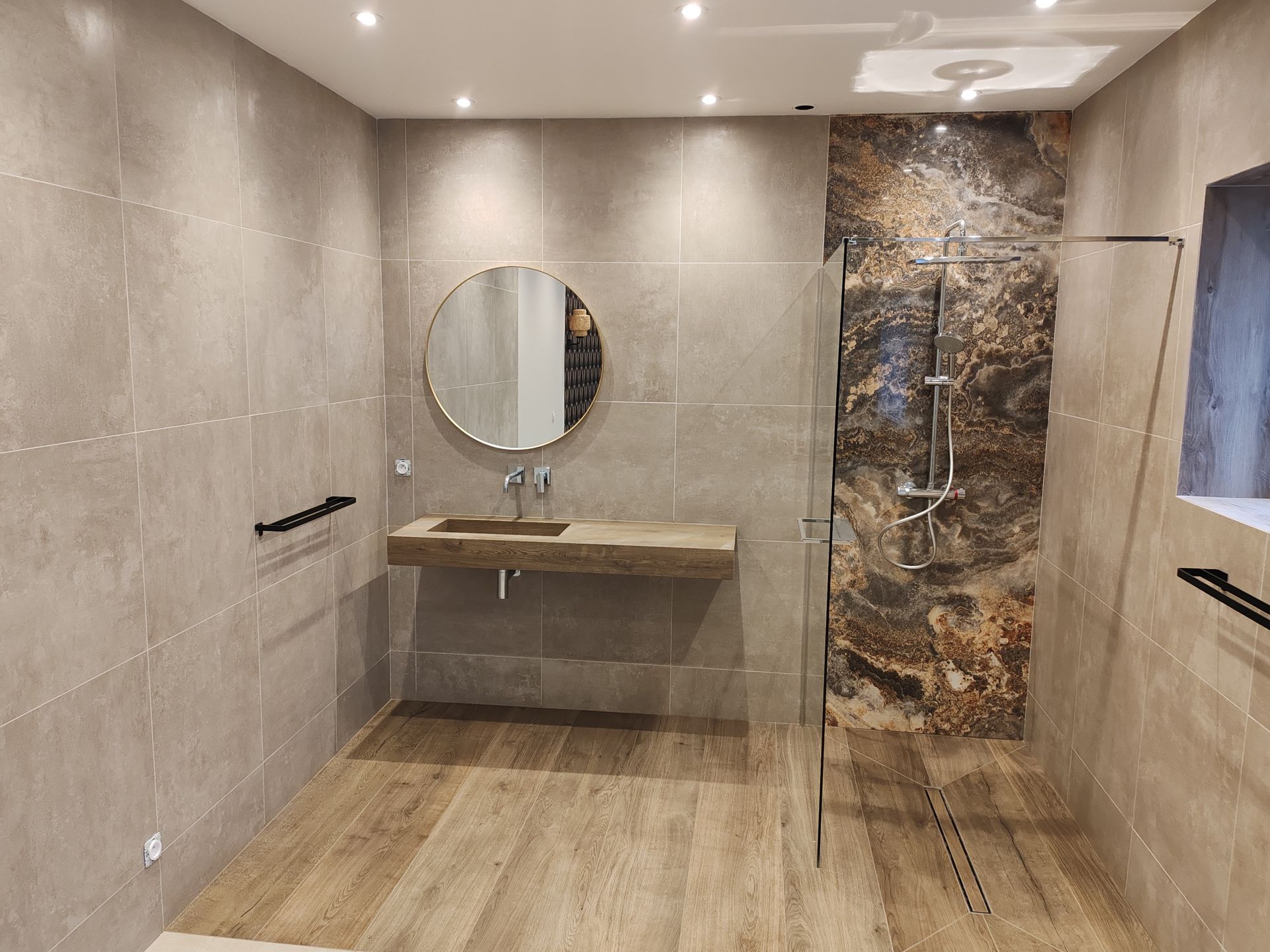 Salle de bains avec carrelage beige, sol imitation bois, miroir rond, lavabo suspendu et douche en verre.