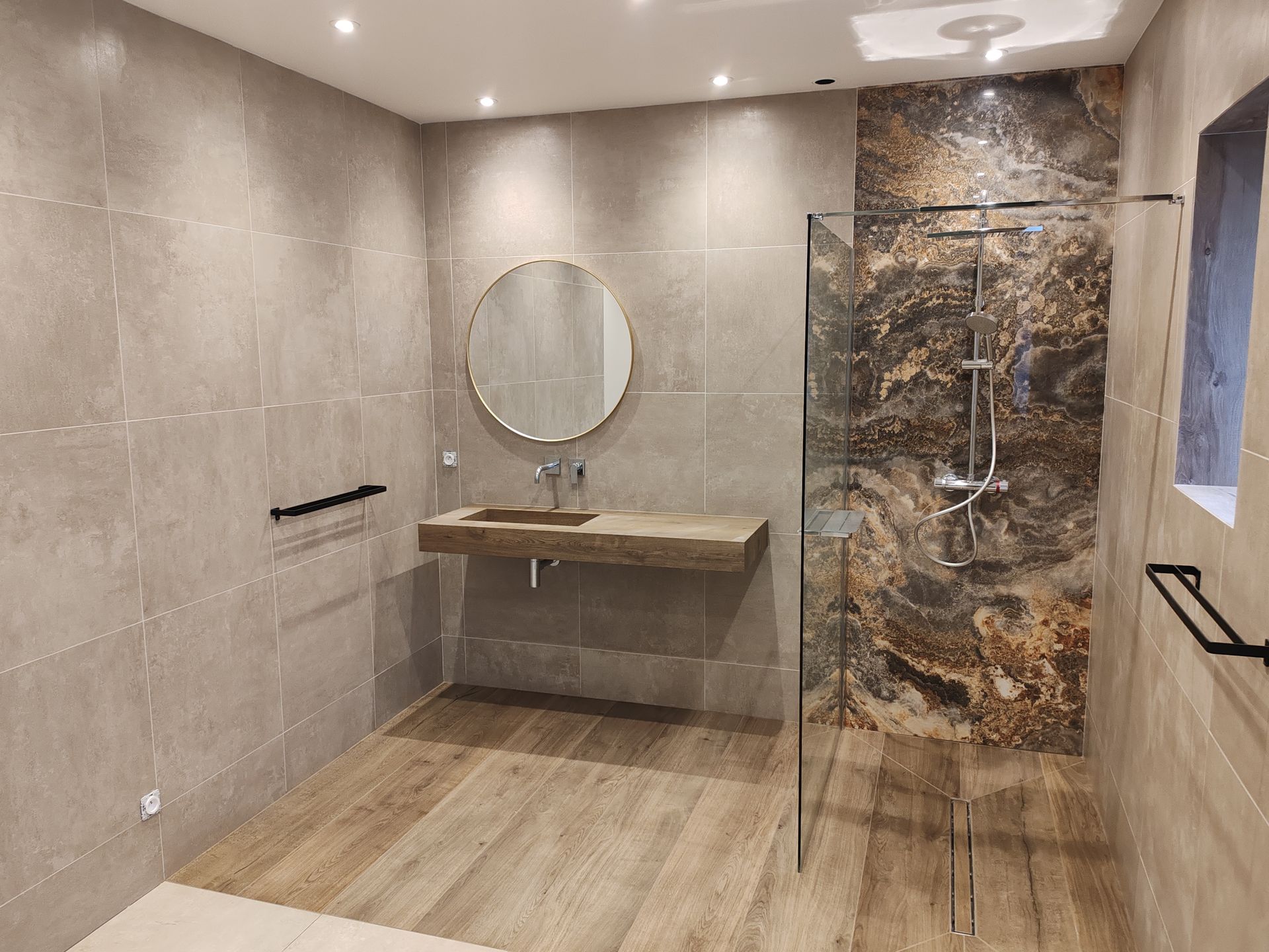 Salle de bains moderne avec sol imitation bois, murs gris, douche en verre et miroir rond au-dessus d'un meuble-lavabo en bois.