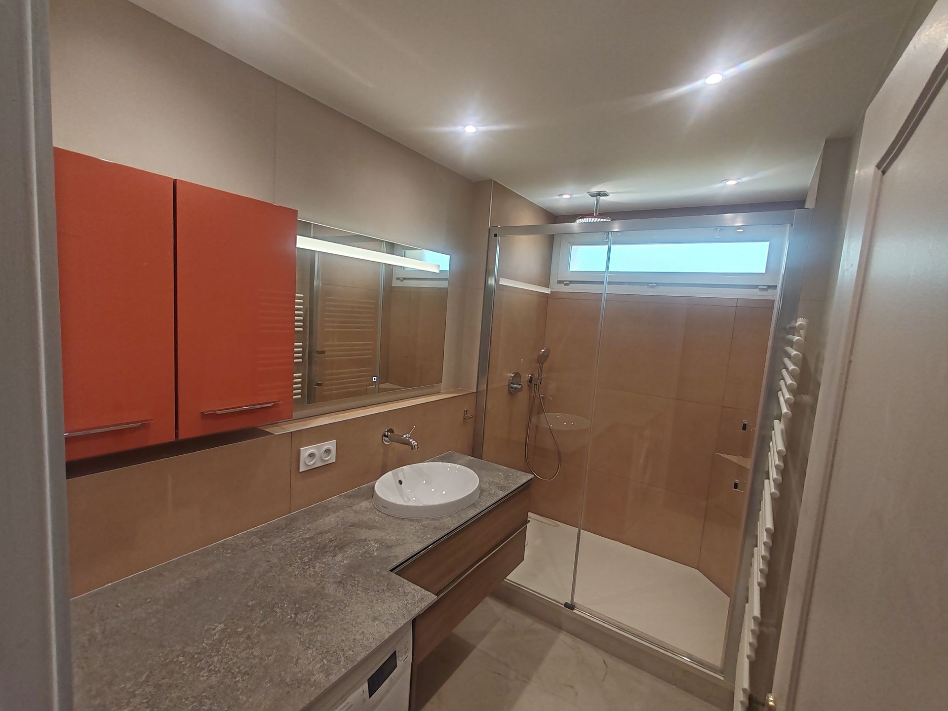 Salle de bains moderne avec meubles orange, comptoir en pierre, lavabo, douche et fenêtre.