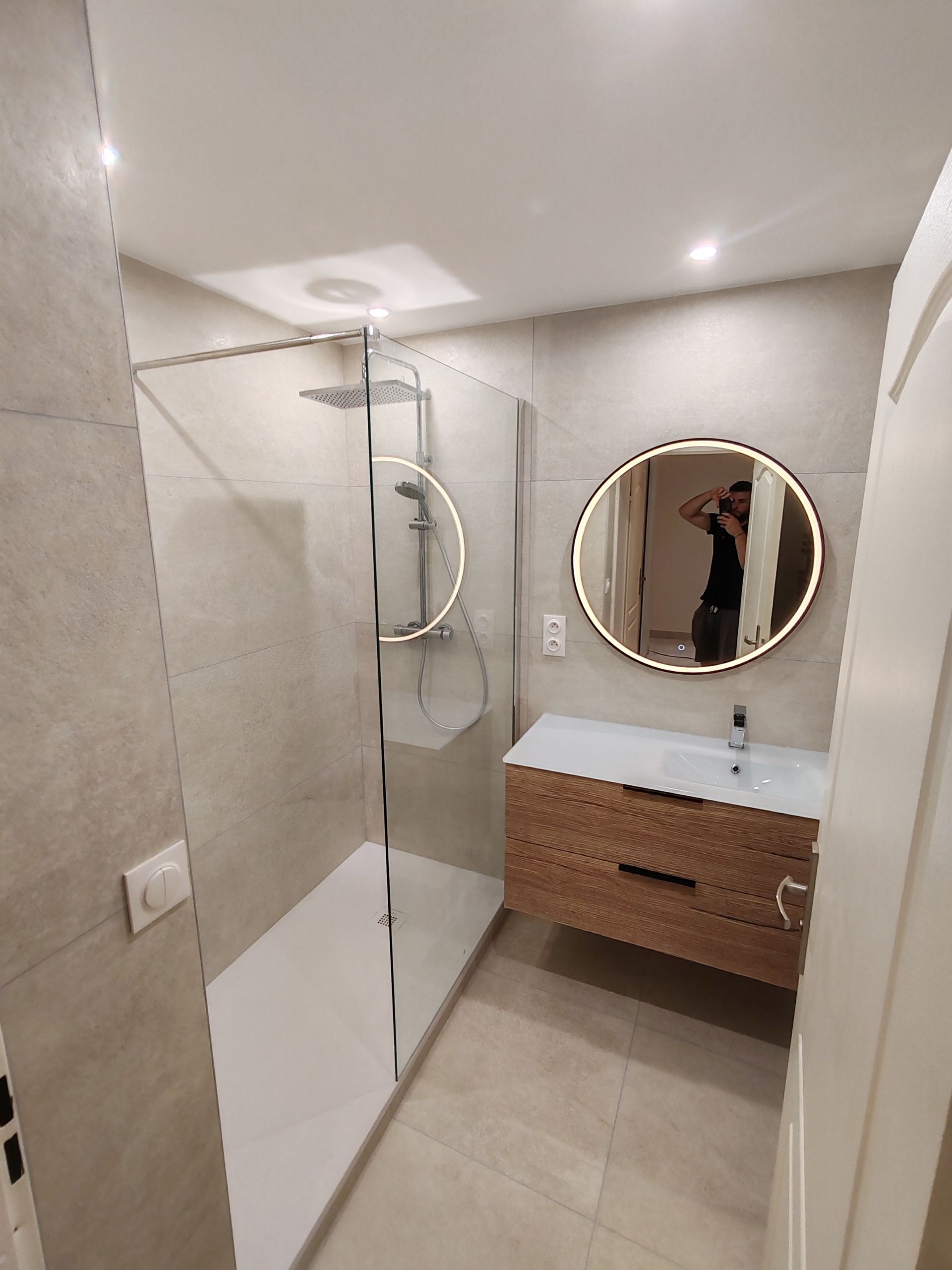 Salle de bains moderne avec douche en verre, meuble-lavabo en bois et miroir rond.