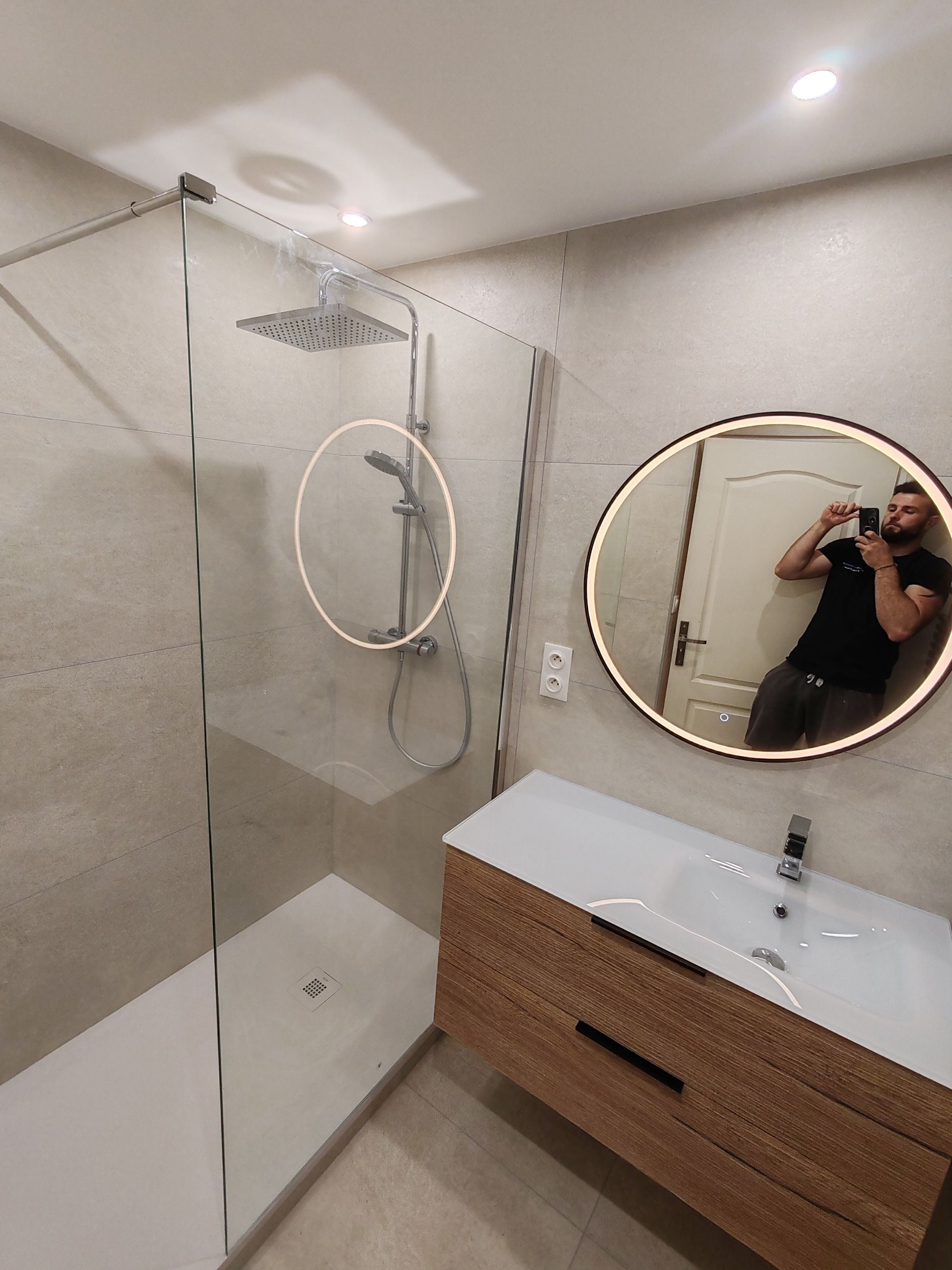 Salle de bains moderne avec douche en verre, lavabo, miroir rond et une personne prenant une photo dans le miroir.