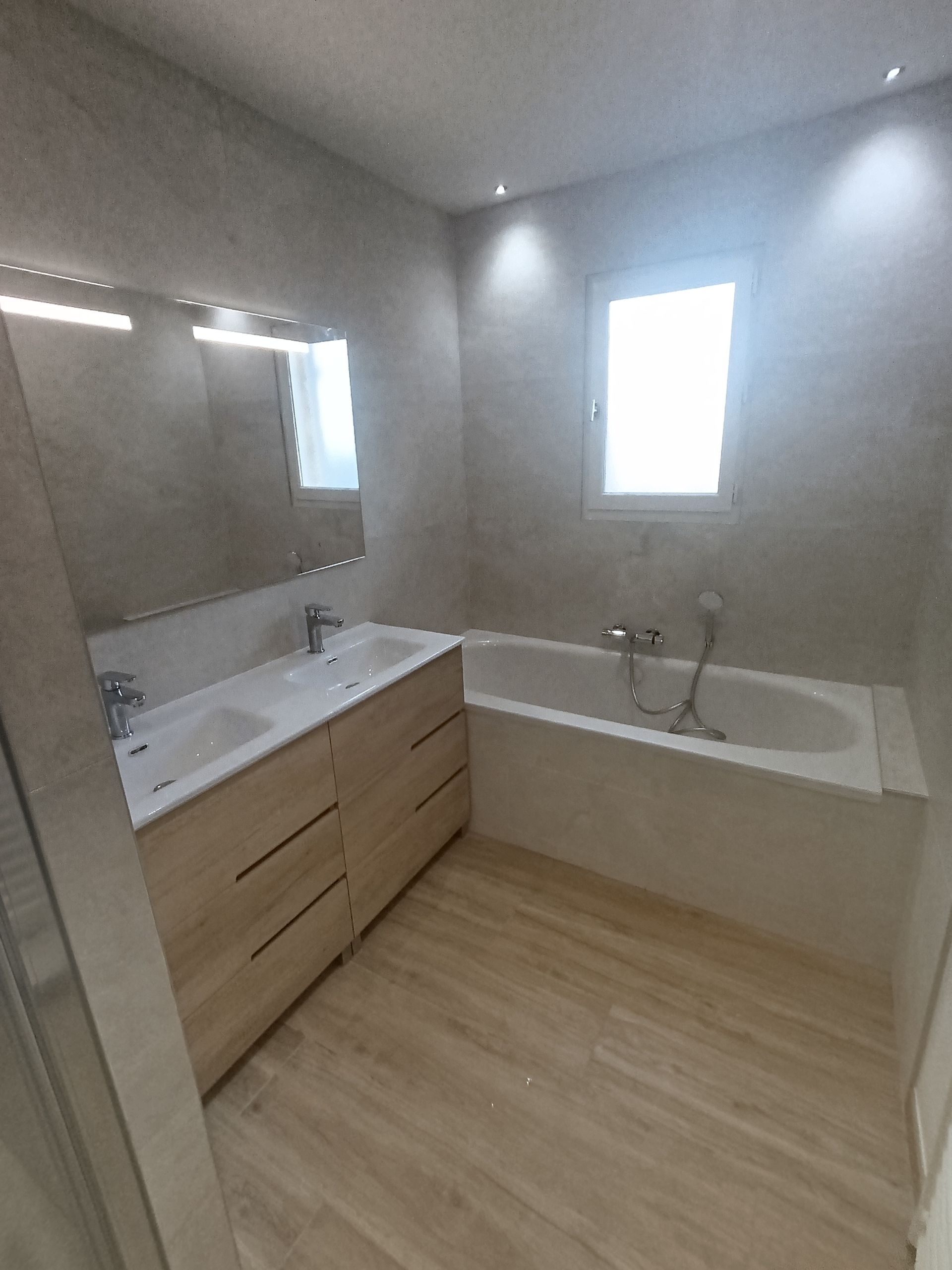 Salle de bains avec meubles en bois clair, baignoire, fenêtre et murs et sol de couleur neutre.