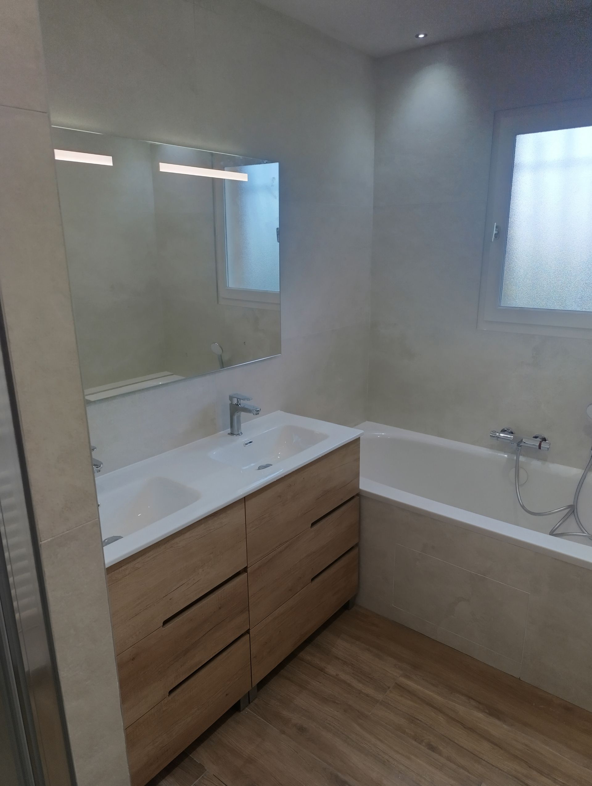 Salle de bains avec meuble-lavabo en bois clair, baignoire et grand miroir. Une fenêtre apporte de la lumière naturelle.