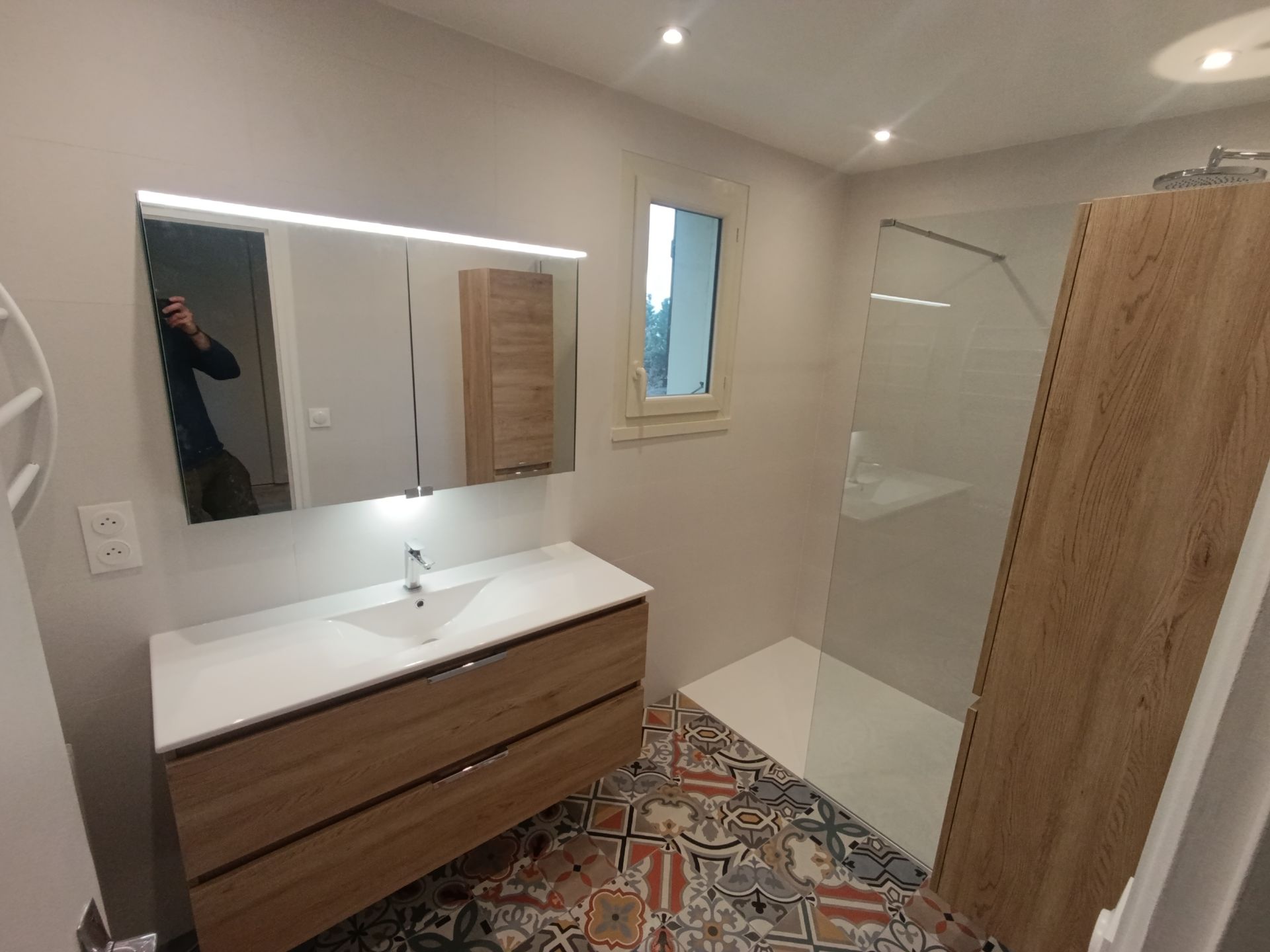 Salle de bains avec meuble-lavabo en bois, grand miroir, douche et carrelage à motifs au sol.