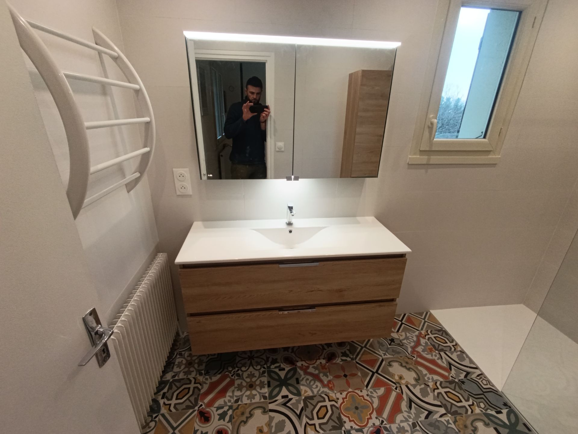 Salle de bains avec meuble-lavabo en bois, sol à motifs, miroir et fenêtre. Un homme se reflète dans le miroir.