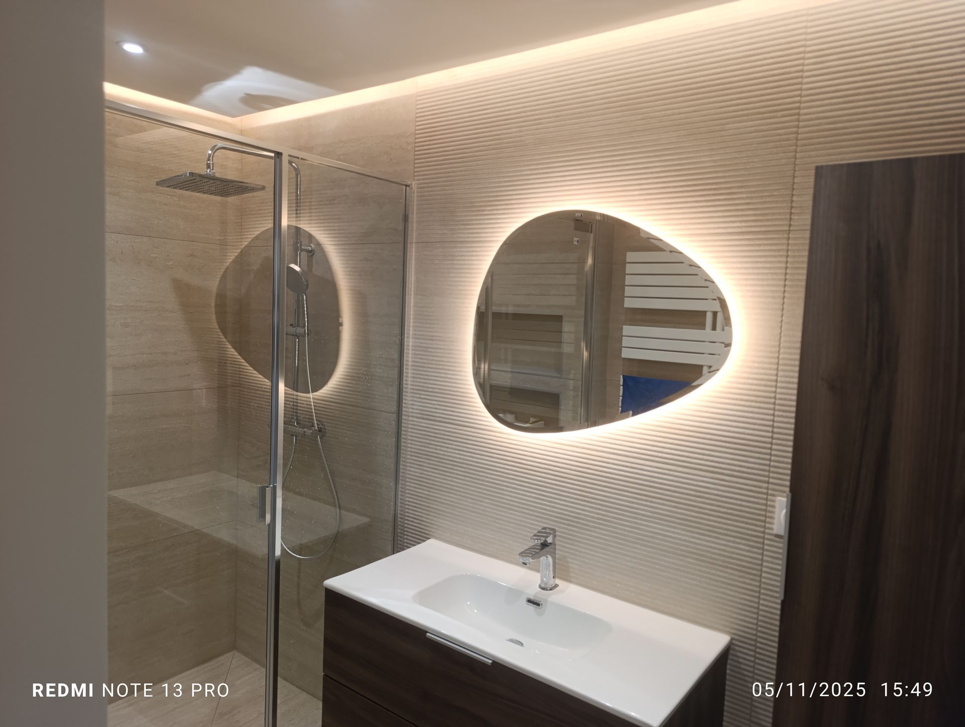 Salle de bains moderne avec mur texturé ondulé, miroirs éclairés, douche en verre et meuble-lavabo.