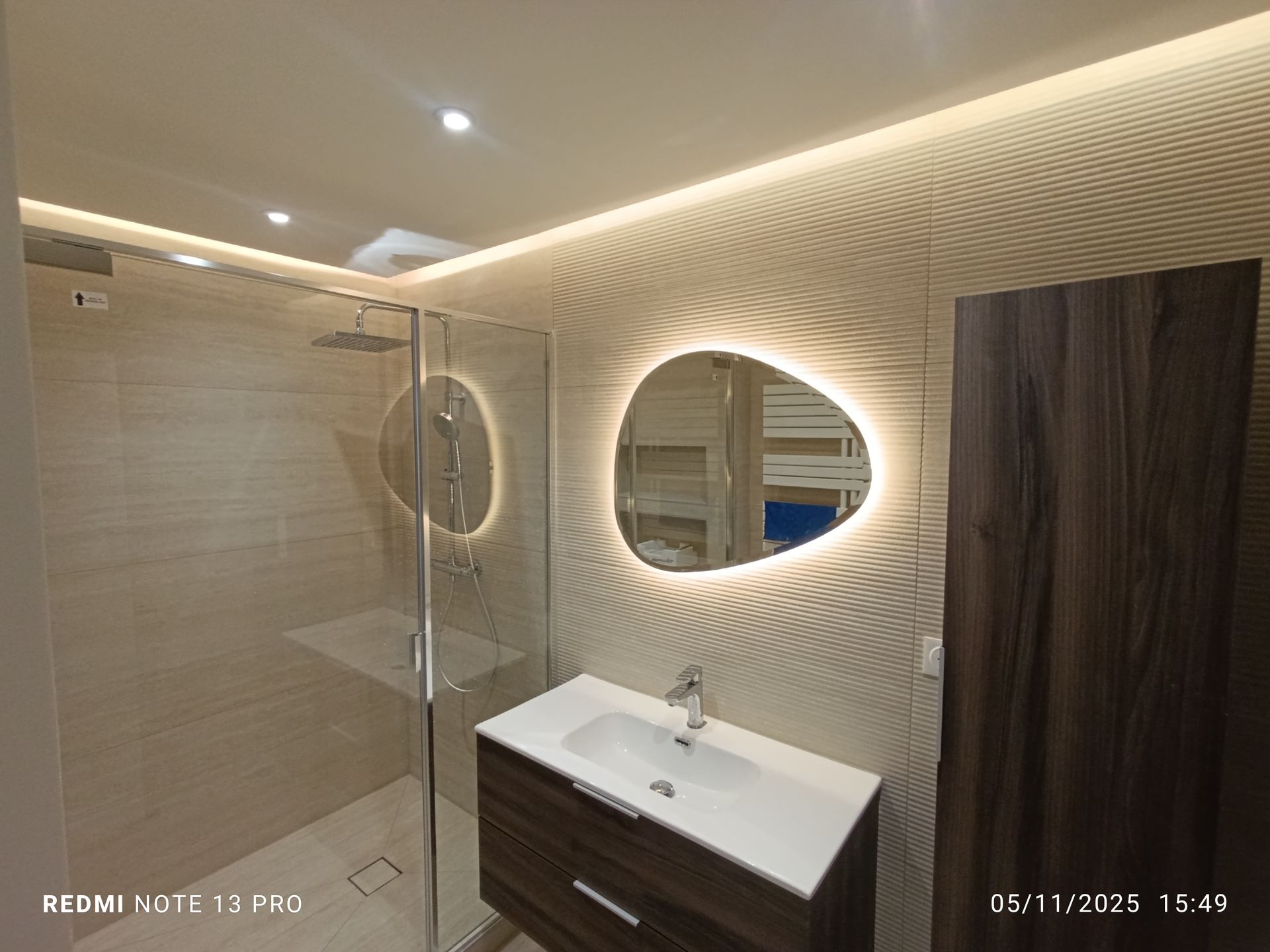 Salle de bains moderne avec miroir éclairé, douche et meuble-lavabo contre un mur texturé.