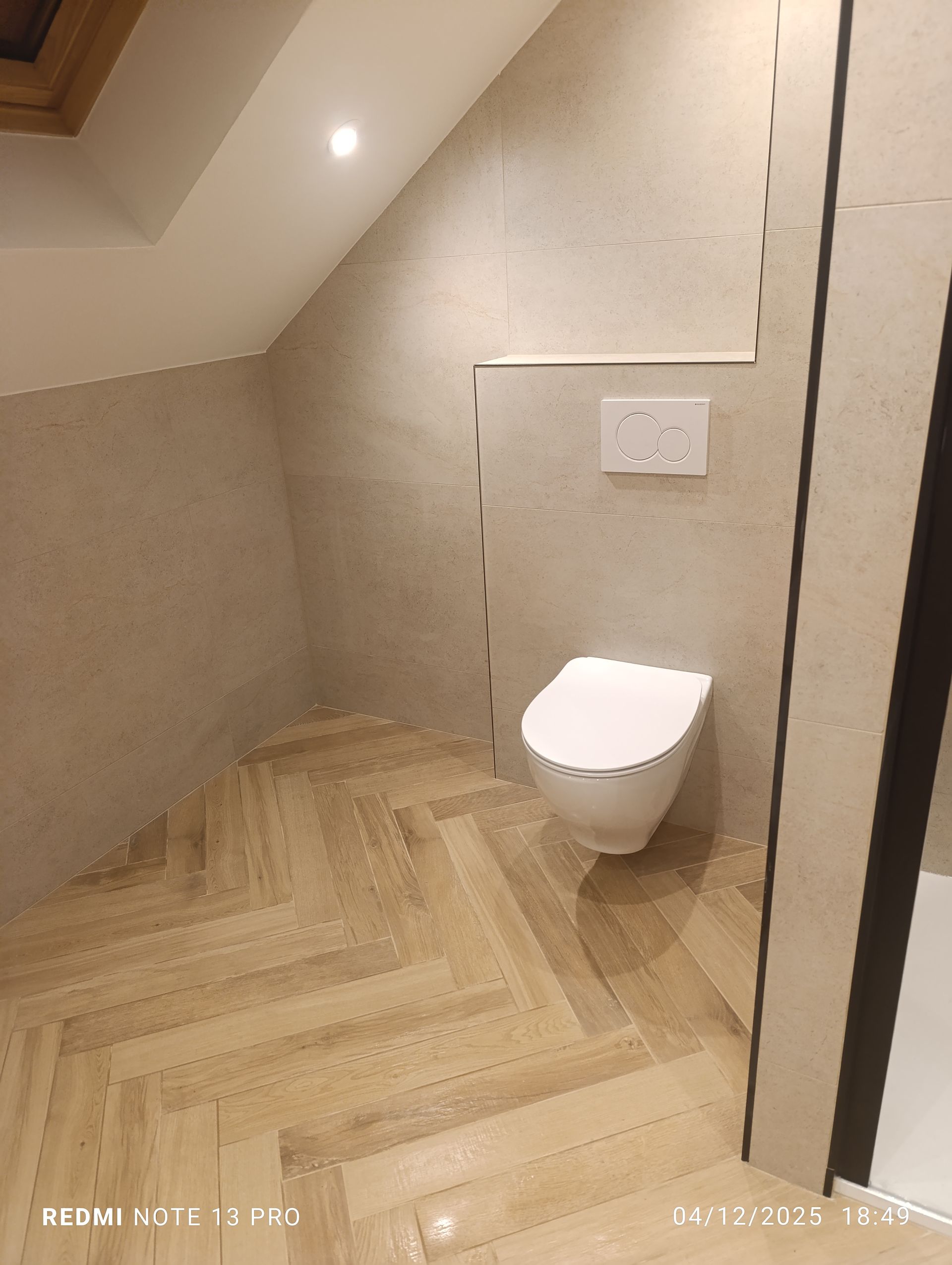 Petite salle de bains avec plafond mansardé, toilettes, parquet clair à chevrons et murs beiges.