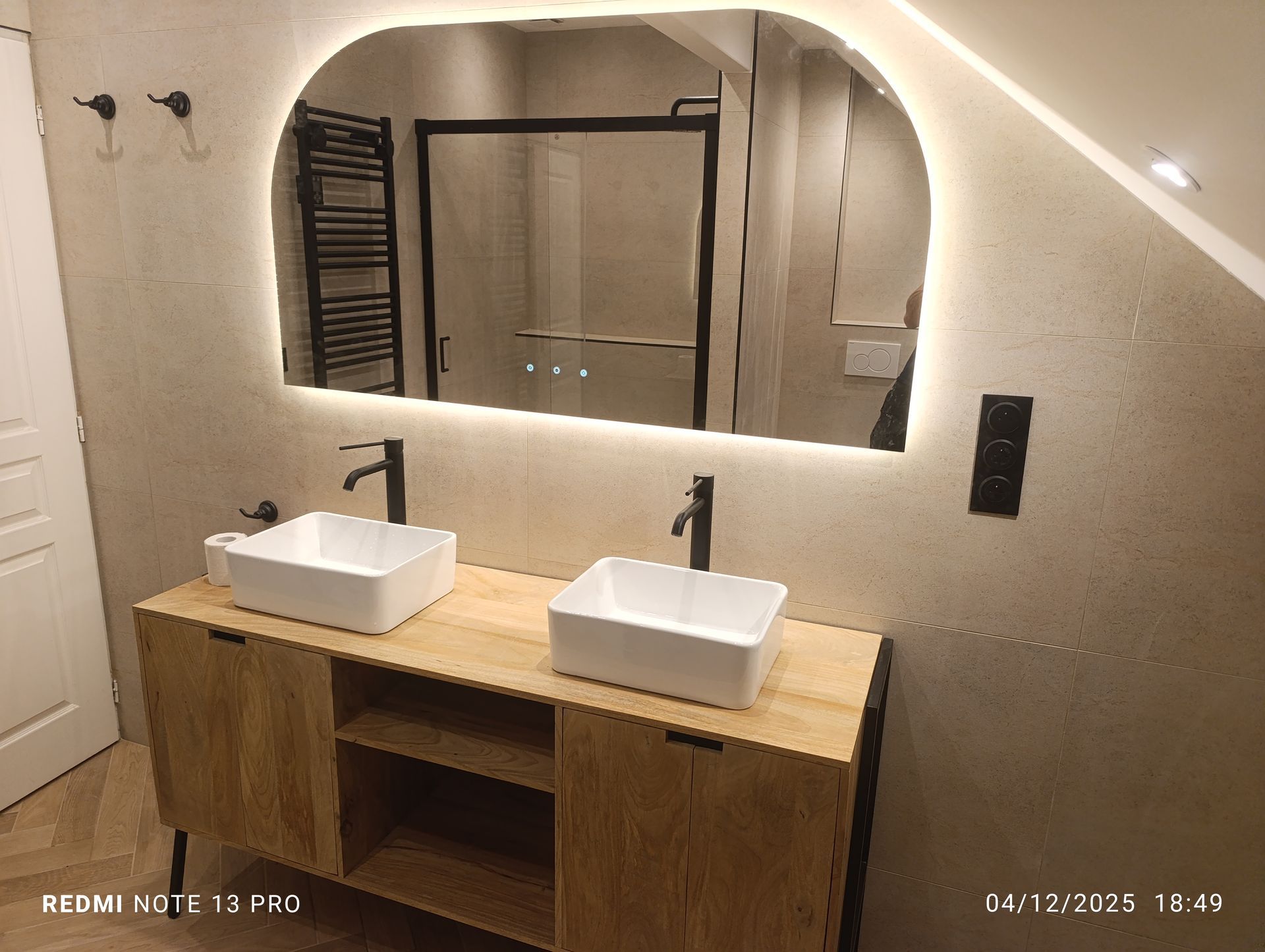 Salle de bains avec double vasque. Grand miroir avec éclairage, deux robinets noirs, meuble en bois, douche à l'arrière.