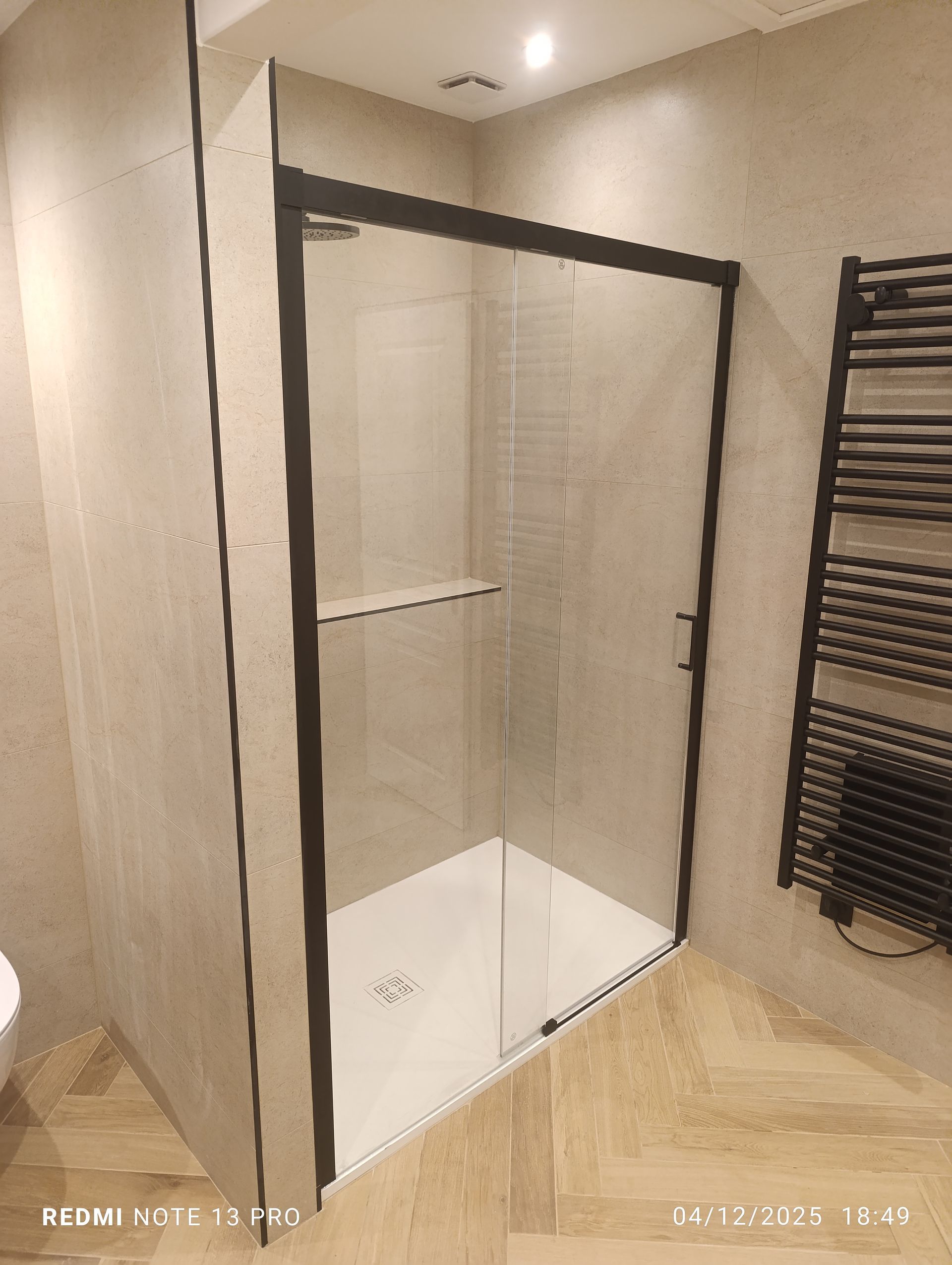 Douche avec cadre noir, porte en verre, receveur blanc et murs carrelés clairs. Un sèche-serviettes noir est placé à droite.