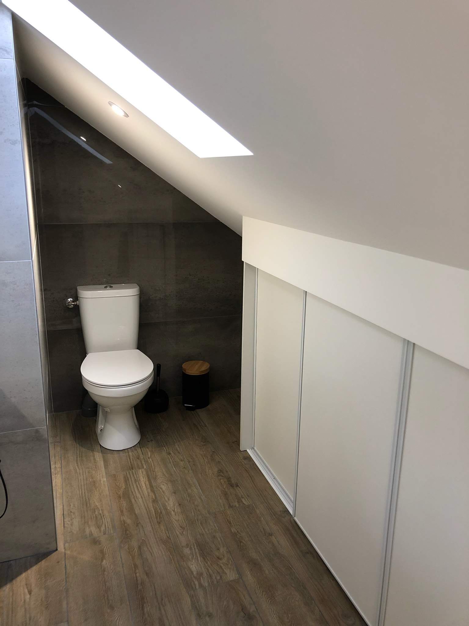 Toilettes dans une petite salle de bains avec plafond mansardé et parquet. Toilettes blanches, murs gris et meubles blancs.