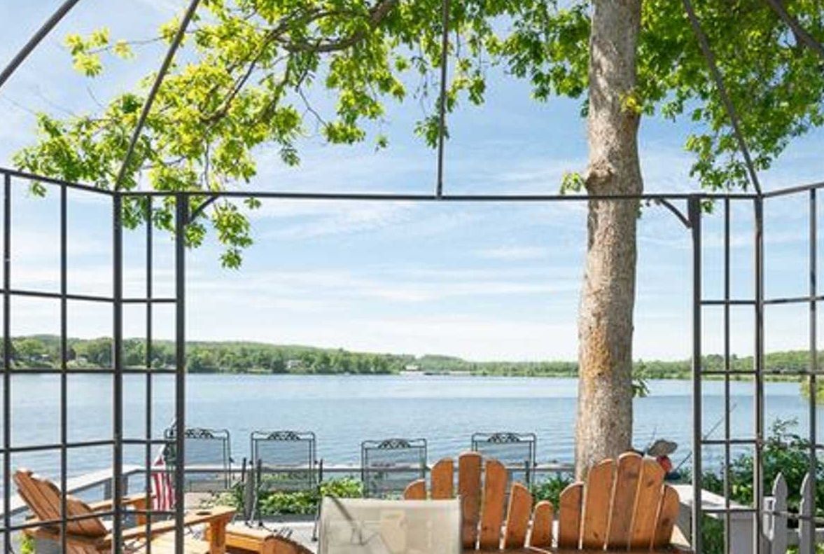 Gazebo with lake view; wooden chairs, tree in center, sunny day.