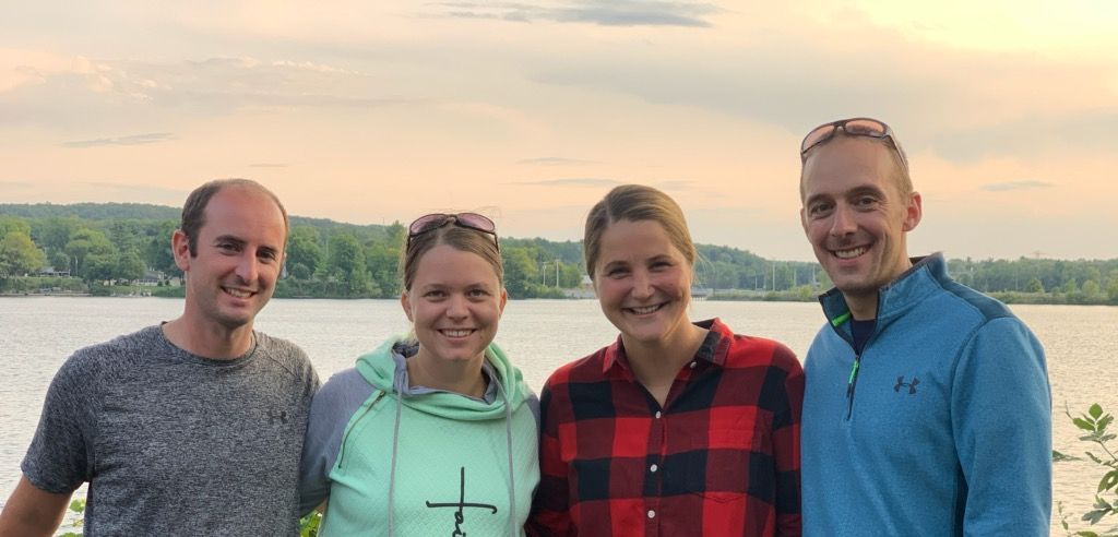 Four people smiling, posing together with a lake and trees in the background. Cloudy sky.
