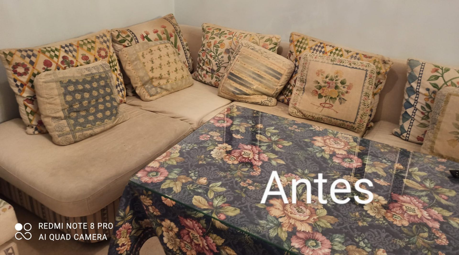Sofá en esquina con cojines estampados y manta floral sobre la mesa; texto “Antes” superpuesto.