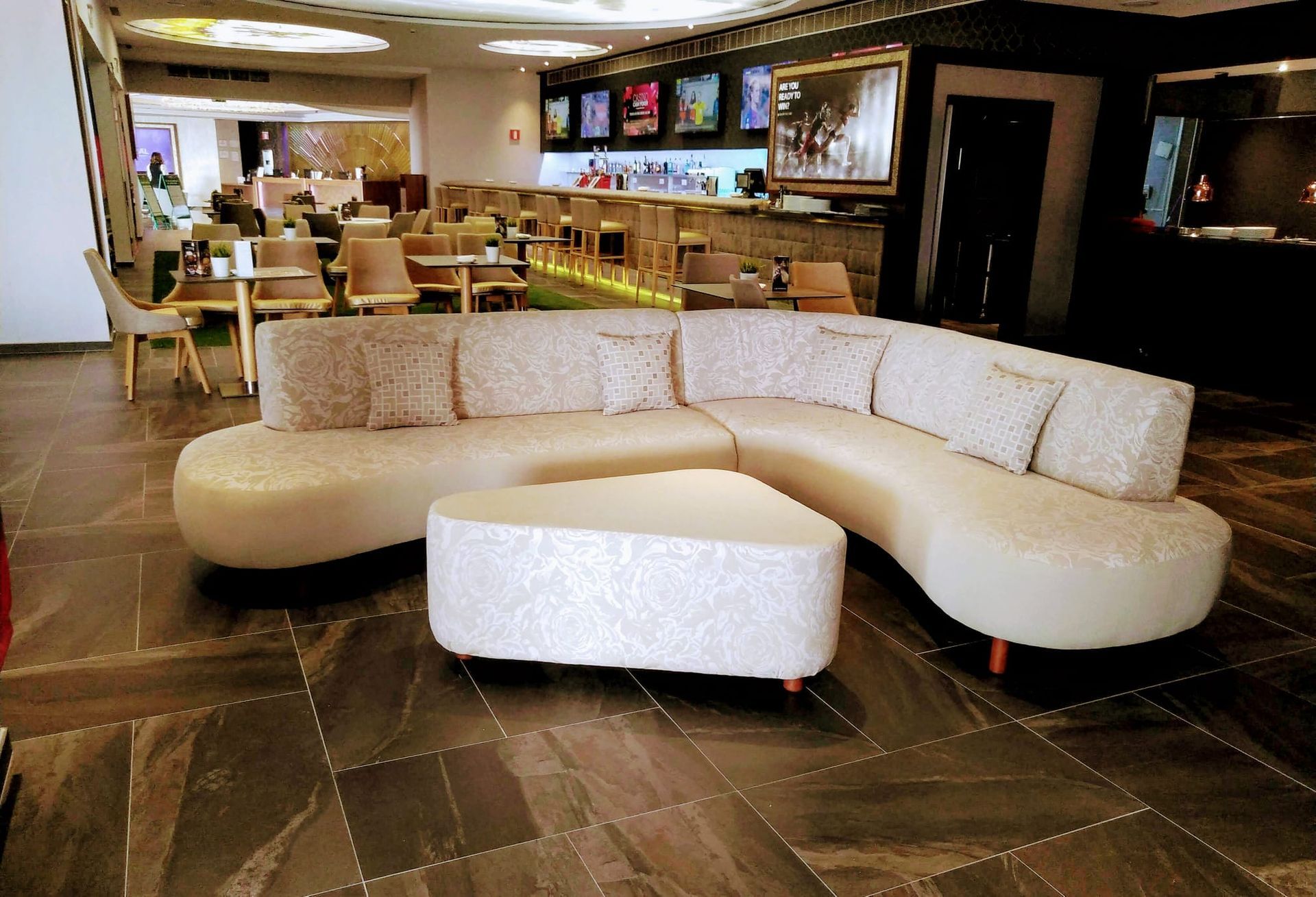 Sofá seccional curvo de color beige en un salón moderno con mesas, sillas y televisores montados en la pared.