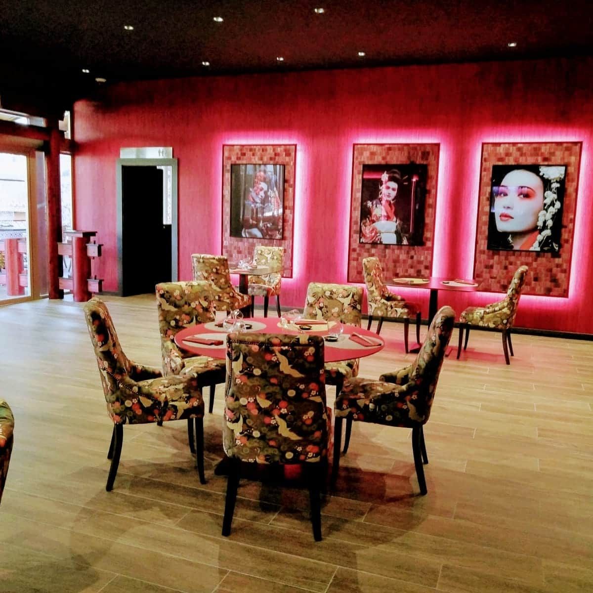 Elegante salón con sillones florales, mesas redondas y paredes iluminadas en rojo que exhiben retratos enmarcados.