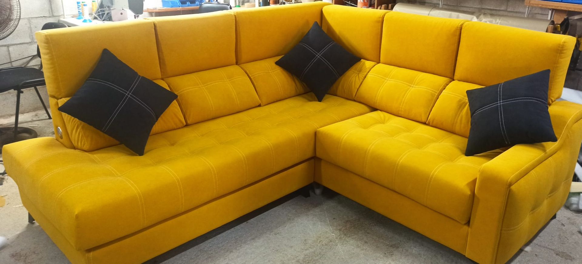 Sofá seccional en forma de L de color amarillo brillante con cojines decorativos negros sobre un suelo gris.